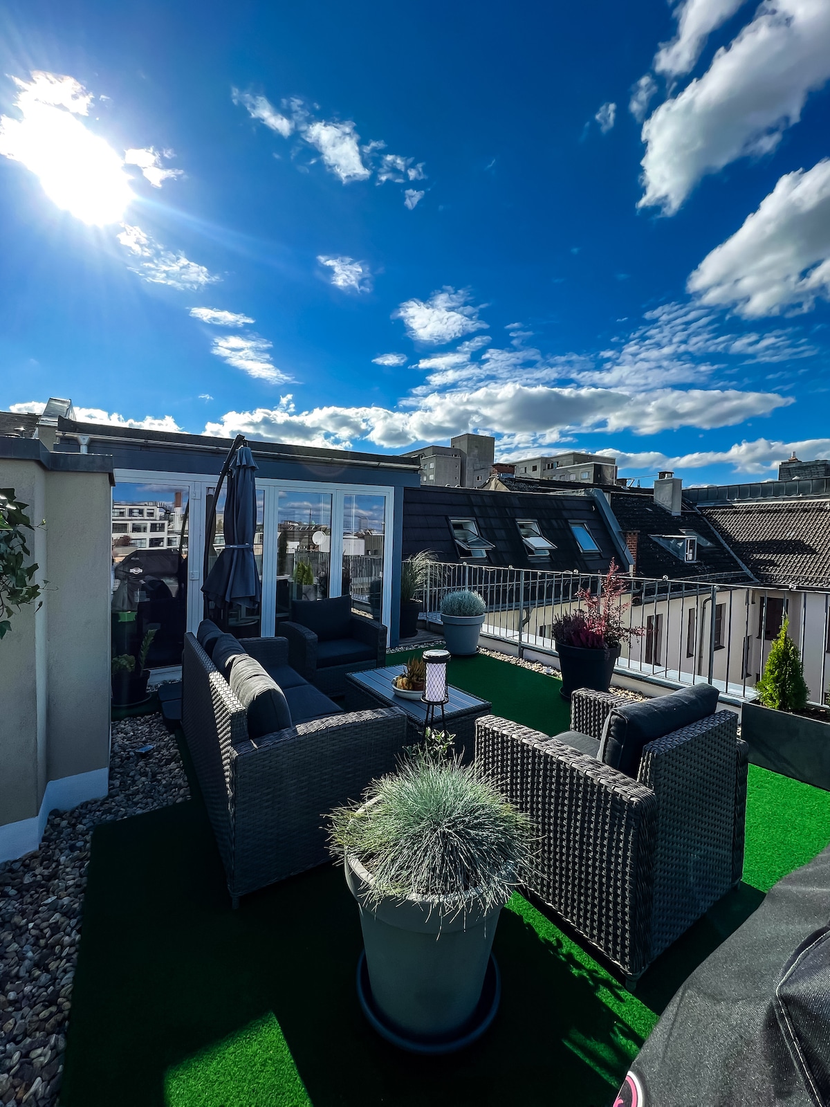 A rooftop terrace is presented, featuring stylish outdoor seating surrounded by potted plants. The area is illuminated by sunlight against a clear blue sky, with city rooftops visible in the background. A cozy yet modern atmosphere is created by the arrangement of furniture.