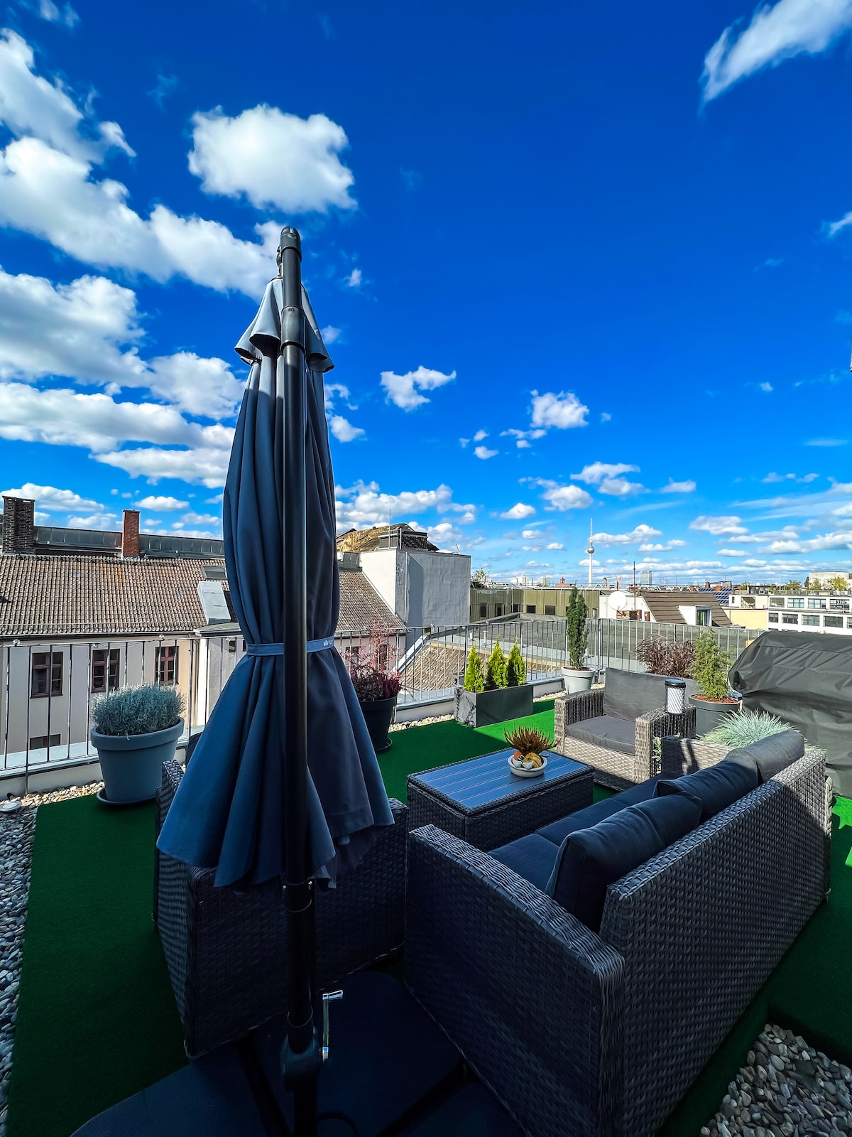 A rooftop terrace is showcased, featuring comfortable seating and a large umbrella for shade. Lush greenery can be seen in planters, and the view includes a vibrant blue sky with scattered clouds. The surrounding architecture reflects a mix of styles, enhancing the urban atmosphere.