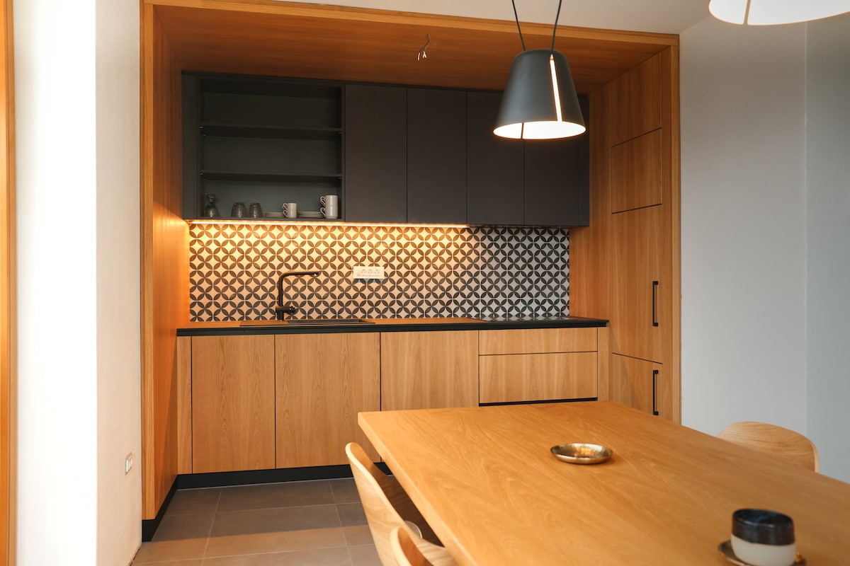 A modern kitchen area is presented, featuring wooden cabinetry and a striking black countertop. Geometric tile backsplashes add visual interest. A wooden dining table with simple chairs is visible, contributing to the functional layout. Pendant lighting casts a warm glow over the space.