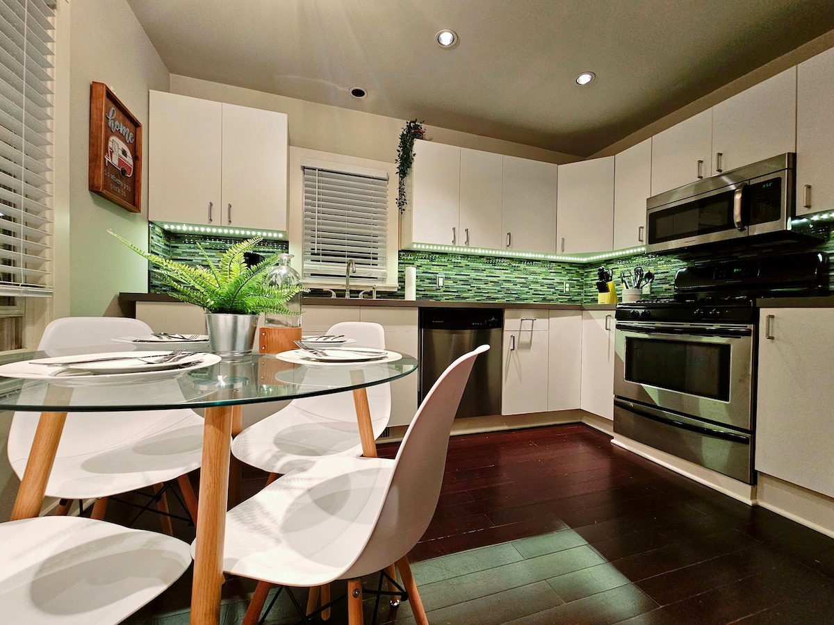 A modern kitchen features sleek, white cabinetry with green accent lighting. A round glass dining table is set with white chairs. Stainless steel appliances, including an oven and a dishwasher, are positioned against a tiled backsplash, enhancing the functional design of the space.