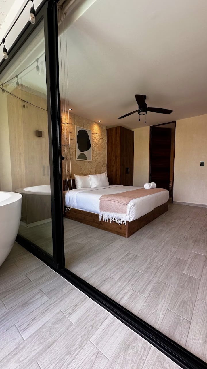 Стая с голямо двойно легло, големи прозорци с изглед към градината и басейна, модерен дизайн с дървени детайли и отделен балкон с вана на открито, идеална за отдих.