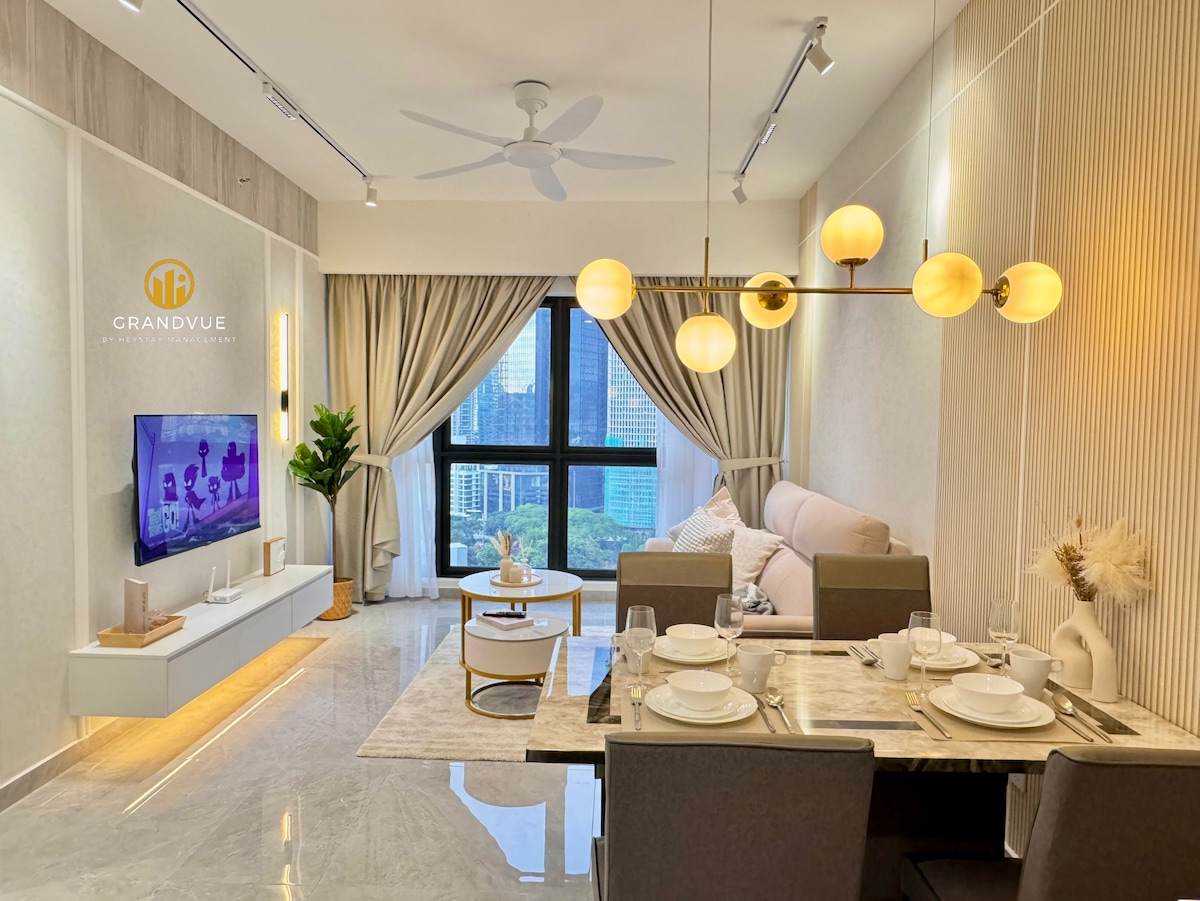 The living area features a light-colored sofa and a coffee table set against a large window offering a city view. A dining table with tableware is positioned nearby, accompanied by a decorative plant. Soft lighting fixtures enhance the space's modern design.