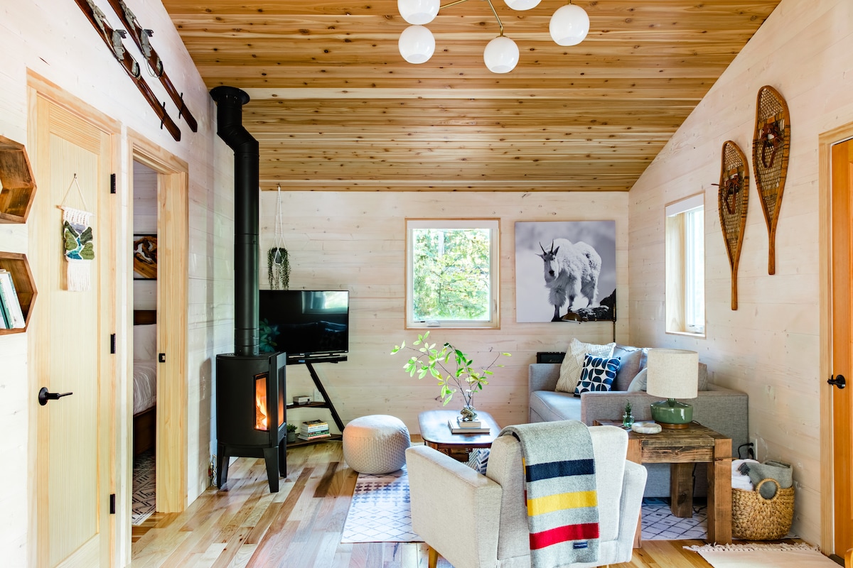 A cozy living area features a gas fireplace, surrounded by natural wood paneling. A comfortable seating arrangement includes a sofa and armchair, with decorative elements like a colorful blanket and a small coffee table. The room is illuminated by modern pendant lights and a window offering scenic views.