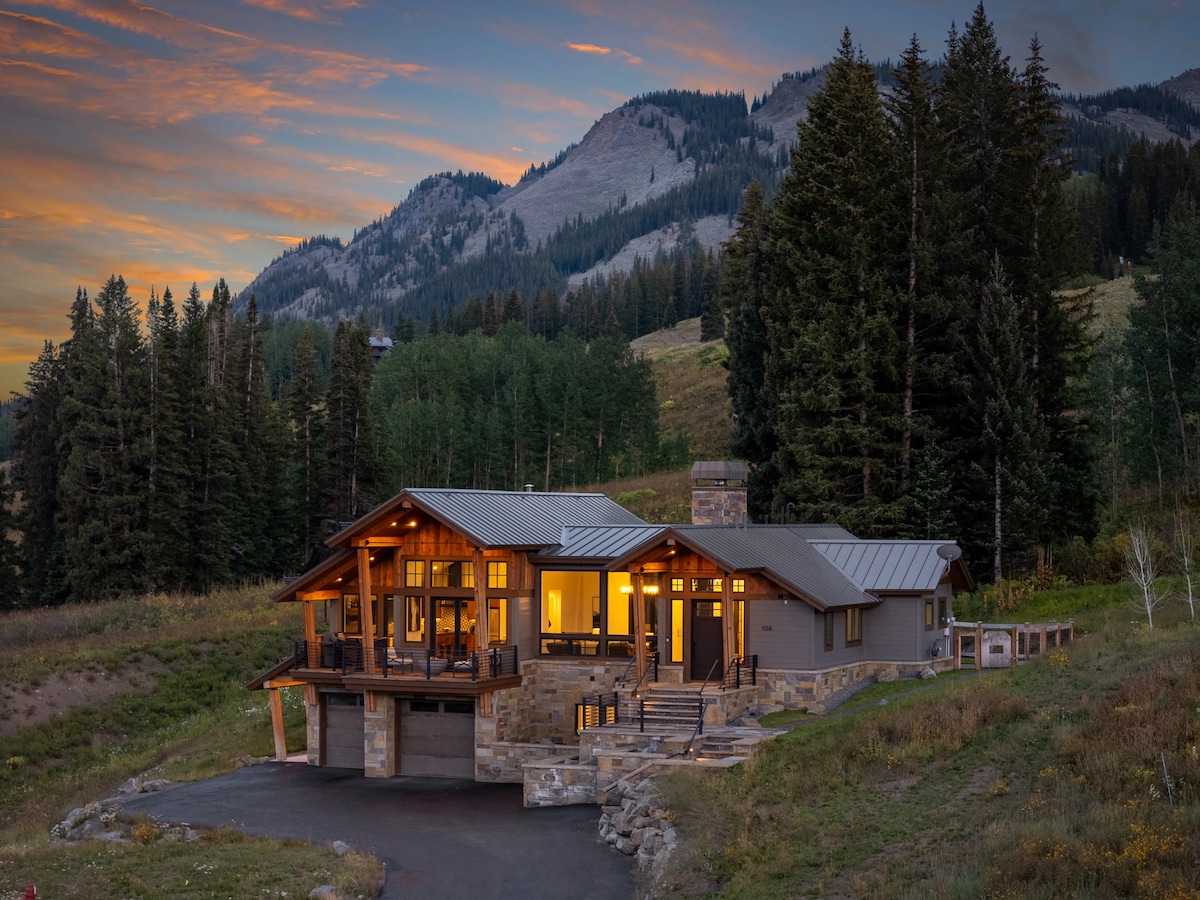 The mountain retreat features a modern exterior with large windows and a spacious deck, surrounded by lush greenery. The backdrop of the majestic mountains and streaked sunset sky enhances the serene setting, creating an inviting atmosphere for guests seeking tranquility.