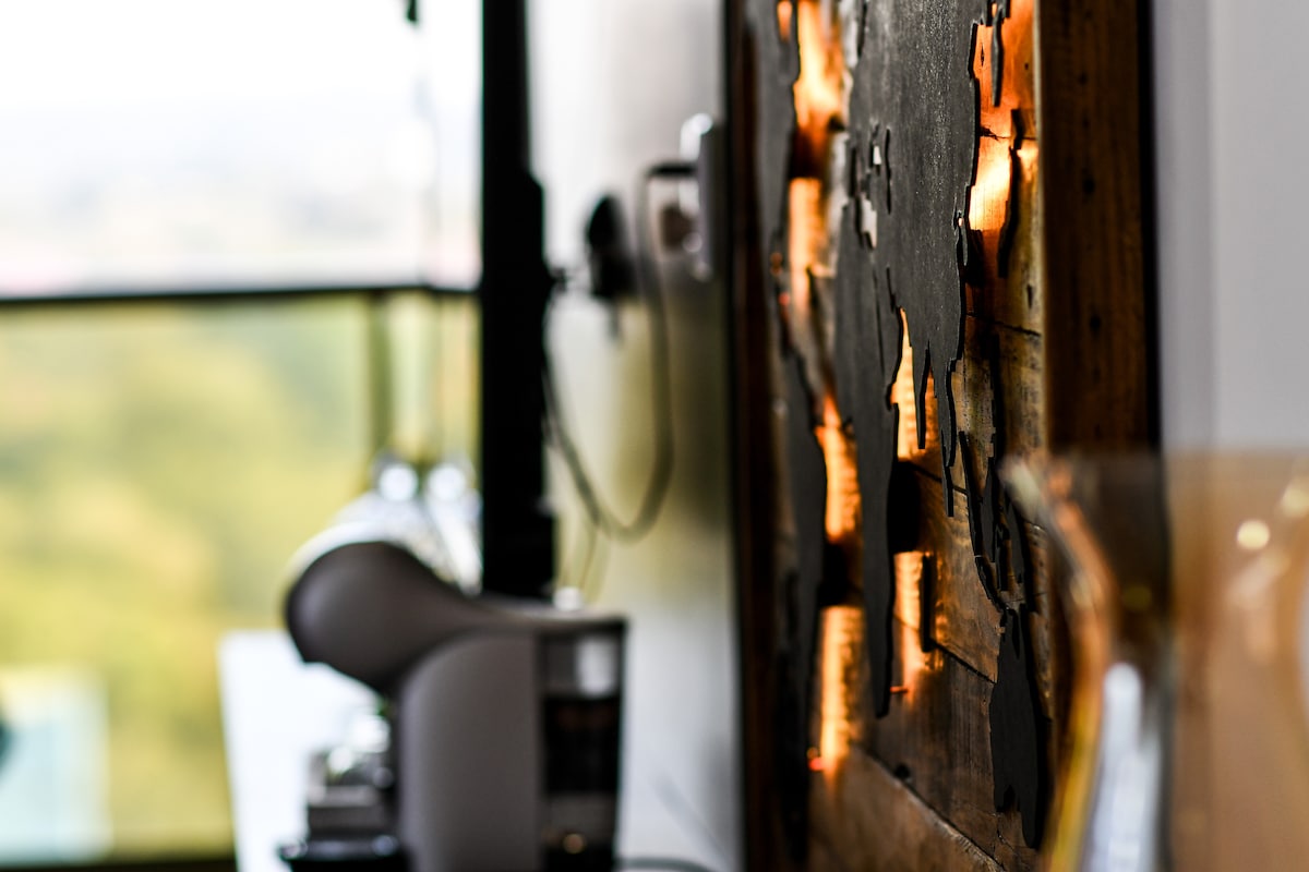 A textured wooden wall features a contemporary map design subtly illuminated, adding a unique decorative touch. The background includes a glimpse of the outdoor greenery through a large window, enhancing the overall ambiance.