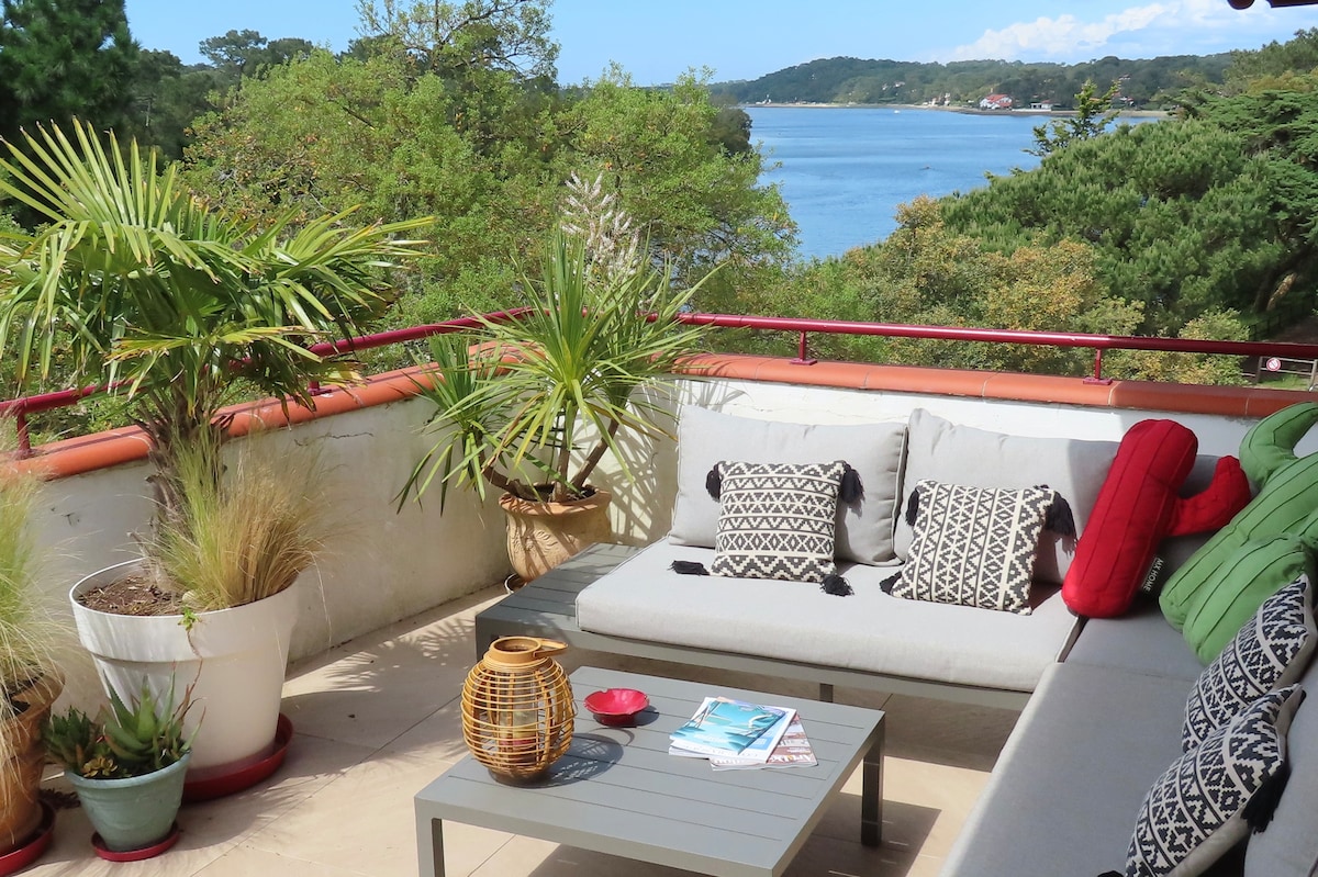 A spacious terrace features a comfortable seating area adorned with patterned cushions. Potted plants add greenery, while a small table holds magazines. The view showcases the serene lake and surrounding trees, creating a peaceful outdoor setting.