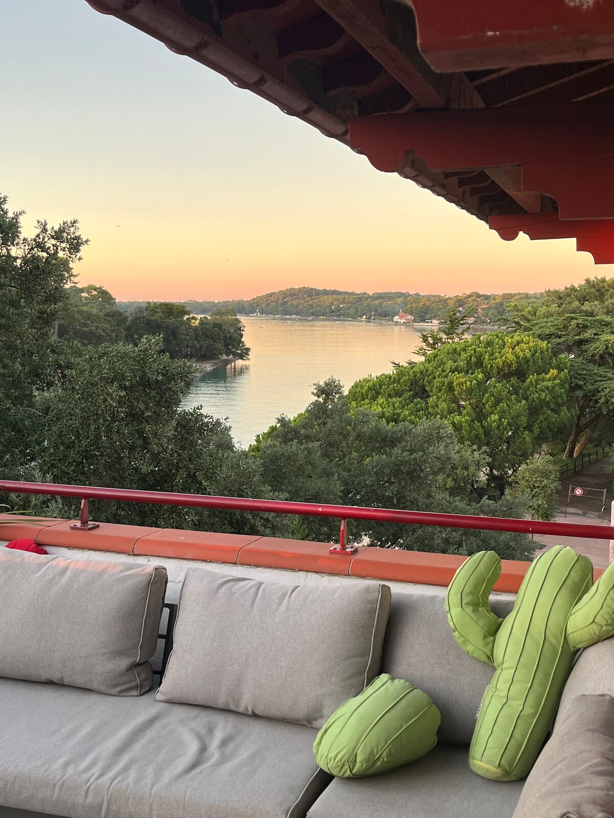 A spacious outdoor seating area is featured, with comfortable gray cushions and decorative green cactus-shaped pillows. In the background, a tranquil waterway reflects soft evening hues, framed by lush greenery and distant hills.