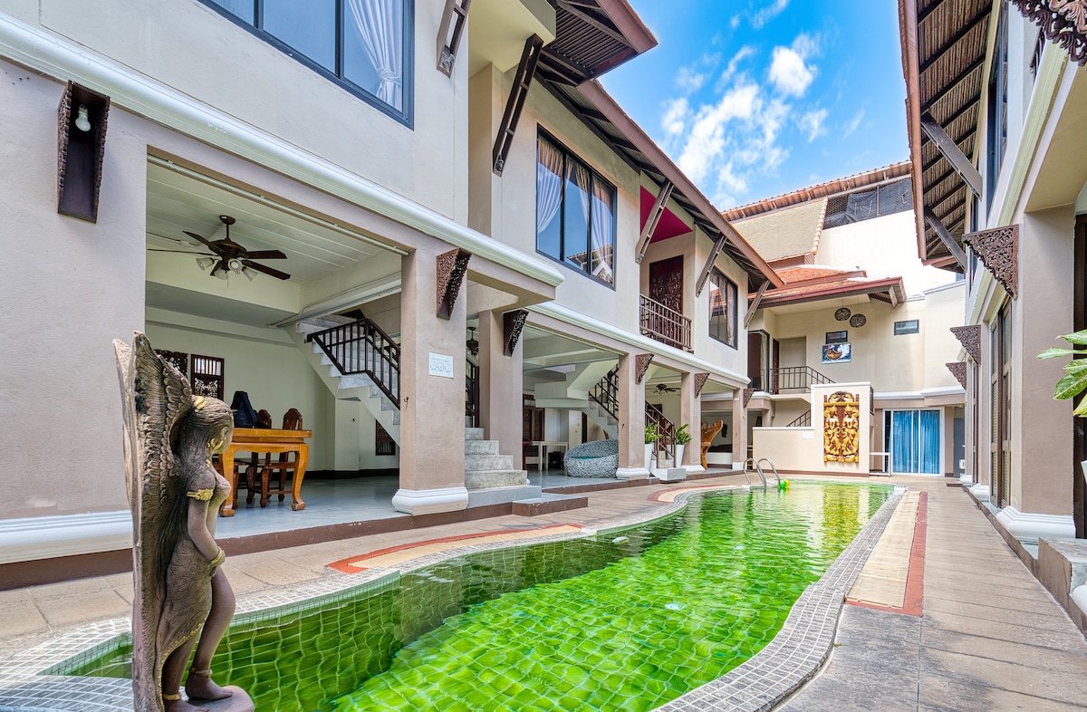 A tranquil free-form swimming pool is surrounded by two-story villa units, showcasing a blend of contemporary architecture and Lanna design. Decorative statues add character to the serene outdoor space, while large windows provide an open view of the inviting interior.