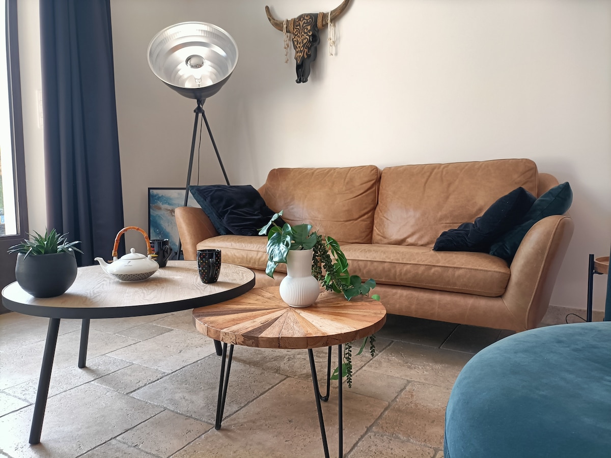 A cozy living area is presented with a brown leather sofa flanked by plush navy cushions. Two coffee tables, one round with a wooden top and another circular, complement the space. A standing lamp and decorative plants add an inviting touch to the room.