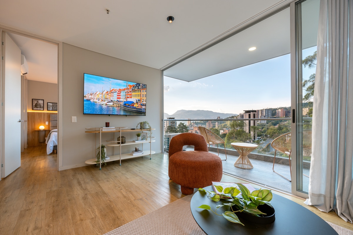 A modern living space features a large smart TV mounted on the wall, complemented by a comfortable orange chair and a coffee table with greenery. Floor-to-ceiling windows offer expansive views of the landscape, allowing natural light to fill the room.