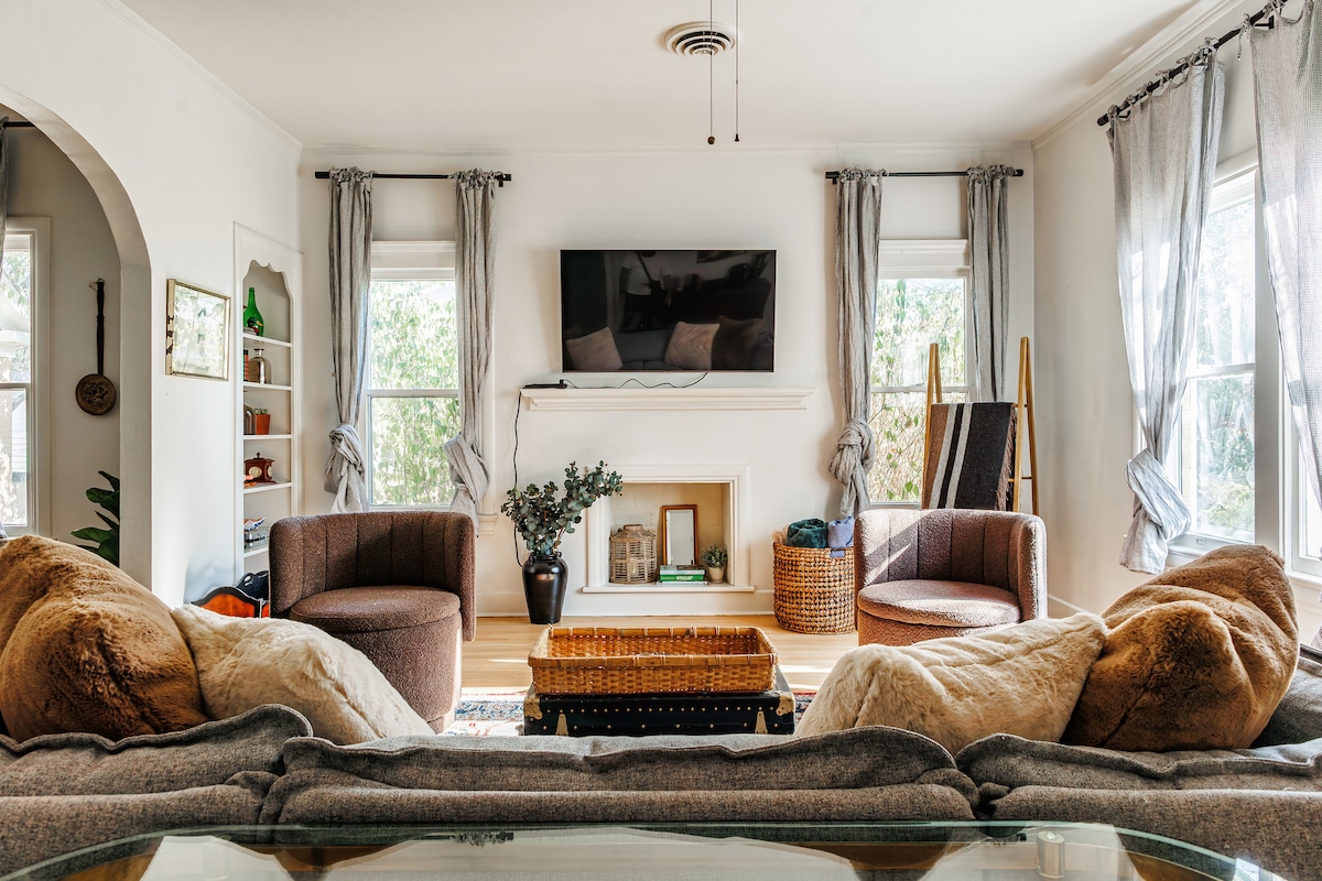 A cozy living room features two armchairs flanking a fireplace, with a large television mounted above the mantel. A soft sofa, adorned with plush pillows, faces a wooden coffee table. Natural light enters through the windows, complemented by sheer curtains and cozy decor.