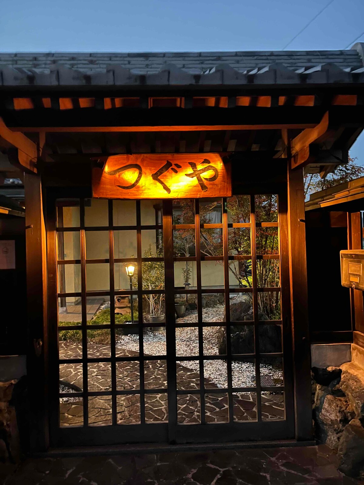 The entrance of the guesthouse is highlighted by a wooden sign bearing the name 'つぐや' illuminated softly. A traditional sliding door with a grid design frames a tranquil garden, featuring stones and softly glowing light fixtures, creating a welcoming ambiance.