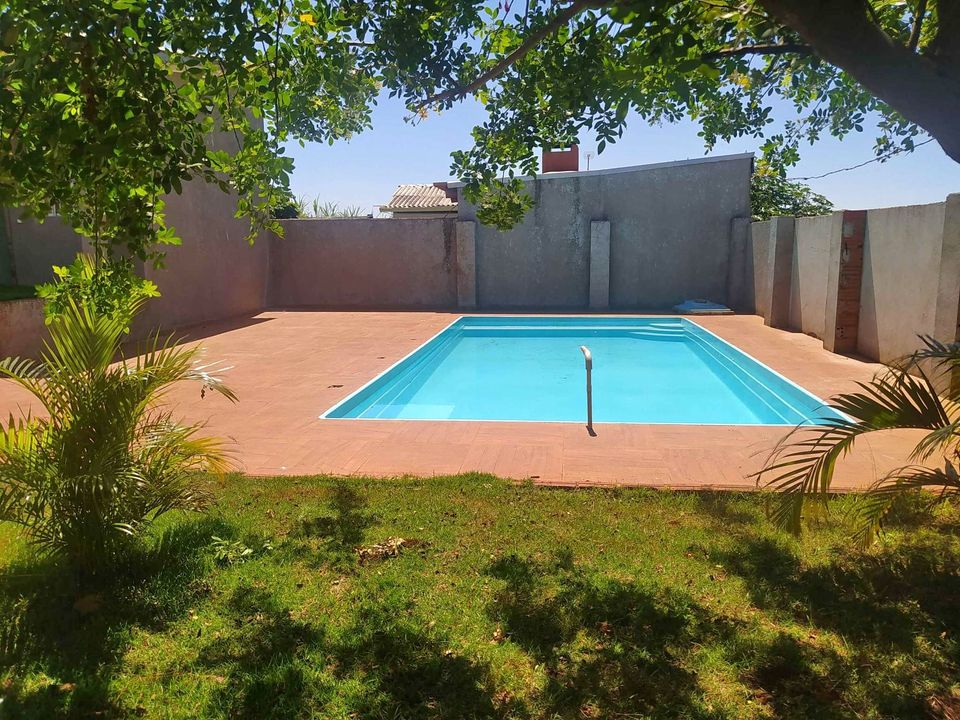 A swimming pool is framed by lush greenery, creating a tranquil environment for relaxation. The pool, set on a tiled surface, features clear blue water and is surrounded by textured walls. Sunlight illuminates the area, enhancing the inviting outdoor space.