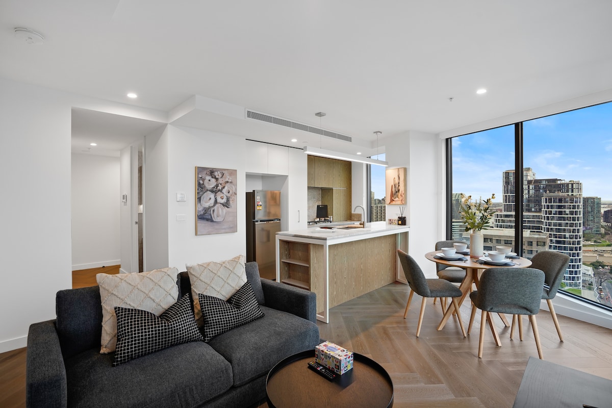 A modern living area features a comfortable sofa adorned with patterned throw pillows. A dining table set for four is positioned nearby, alongside a sleek kitchen with streamlined cabinetry and a large window offering views of the city skyline.