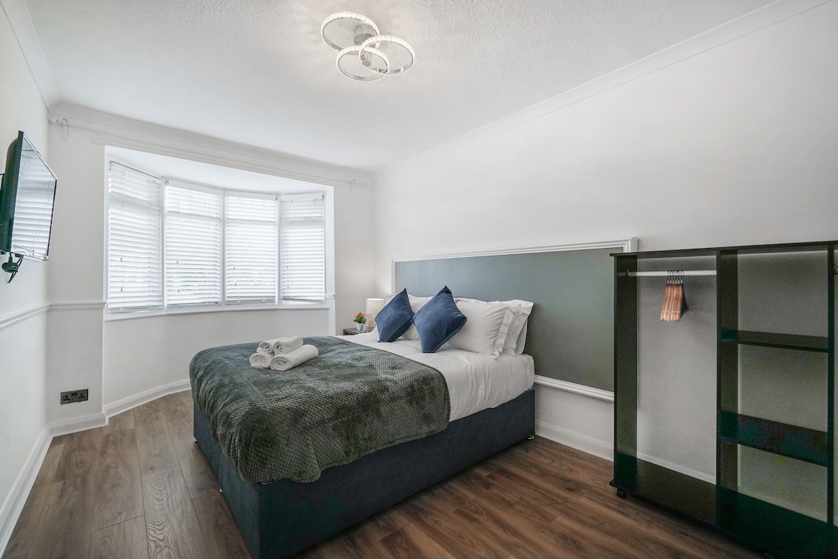 A well-lit bedroom is presented with a spacious bed featuring a dark blue base and plush pillows in shades of blue. A large bay window allows natural light to fill the room, complemented by sleek blinds. A modern green storage unit and a wall-mounted television are visible.