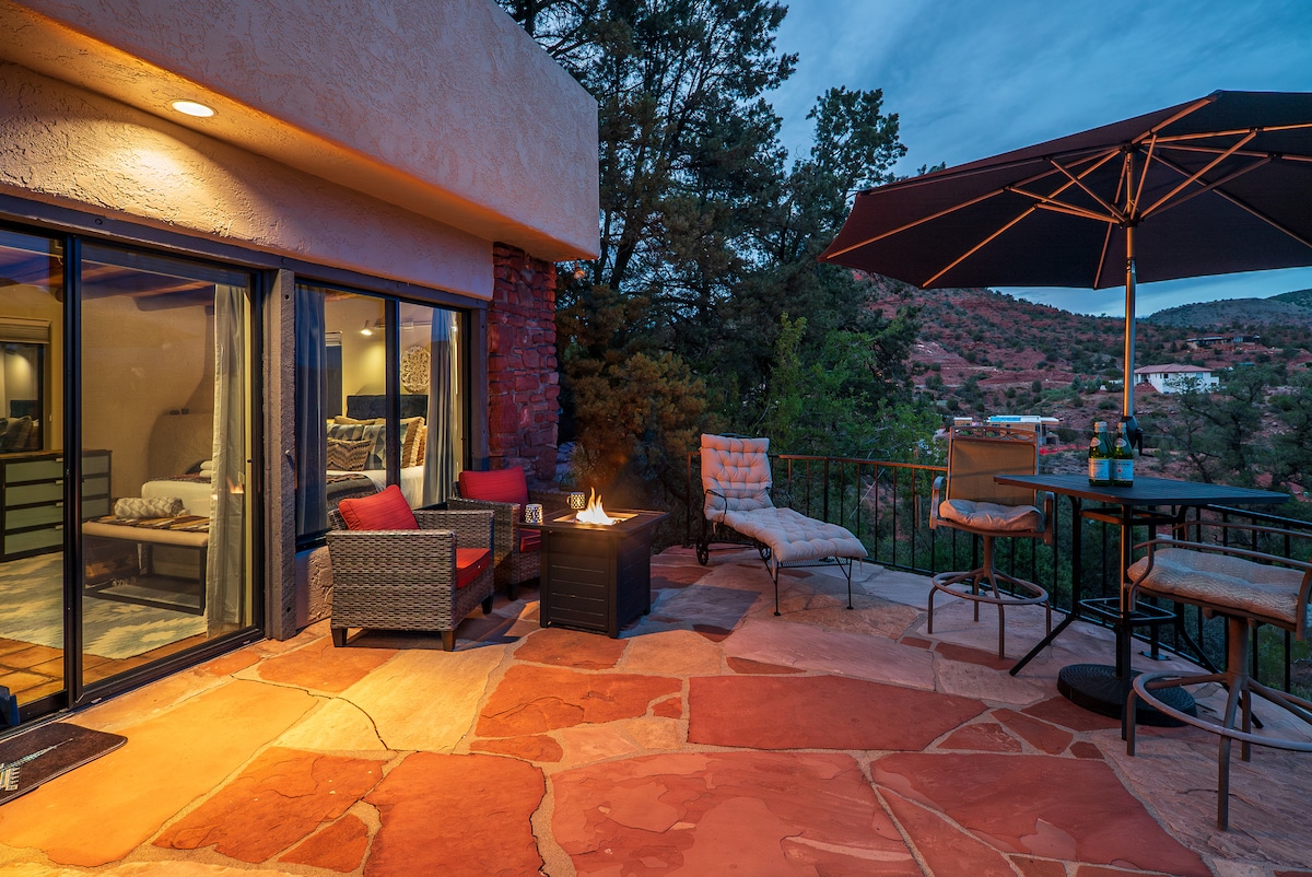 An outdoor terrace features cozy lounge seating and a gas fire pit surrounded by natural stone flooring. A patio umbrella provides shade over the seating area, while views of the lush greenery and red rock formations are visible in the background.