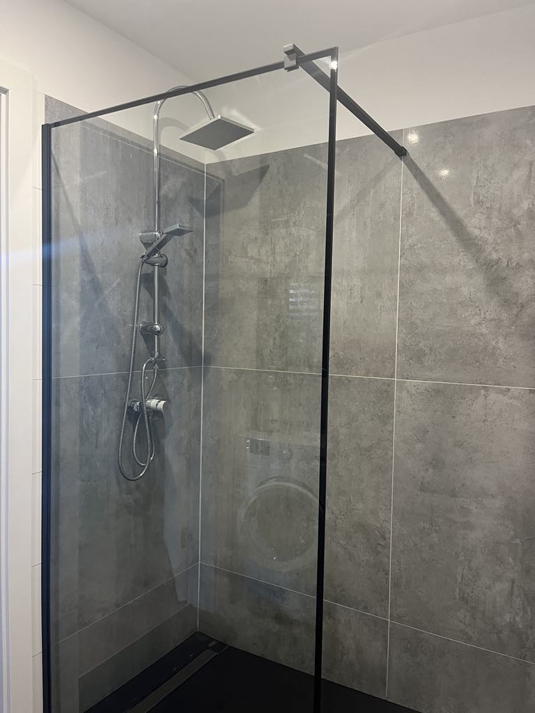 A walk-in shower features sleek gray tile walls and a glass enclosure. A modern showerhead and handheld fixture are mounted on the wall, providing a contemporary look. A washing machine is visible behind the glass panel, integrating functionality within the space.