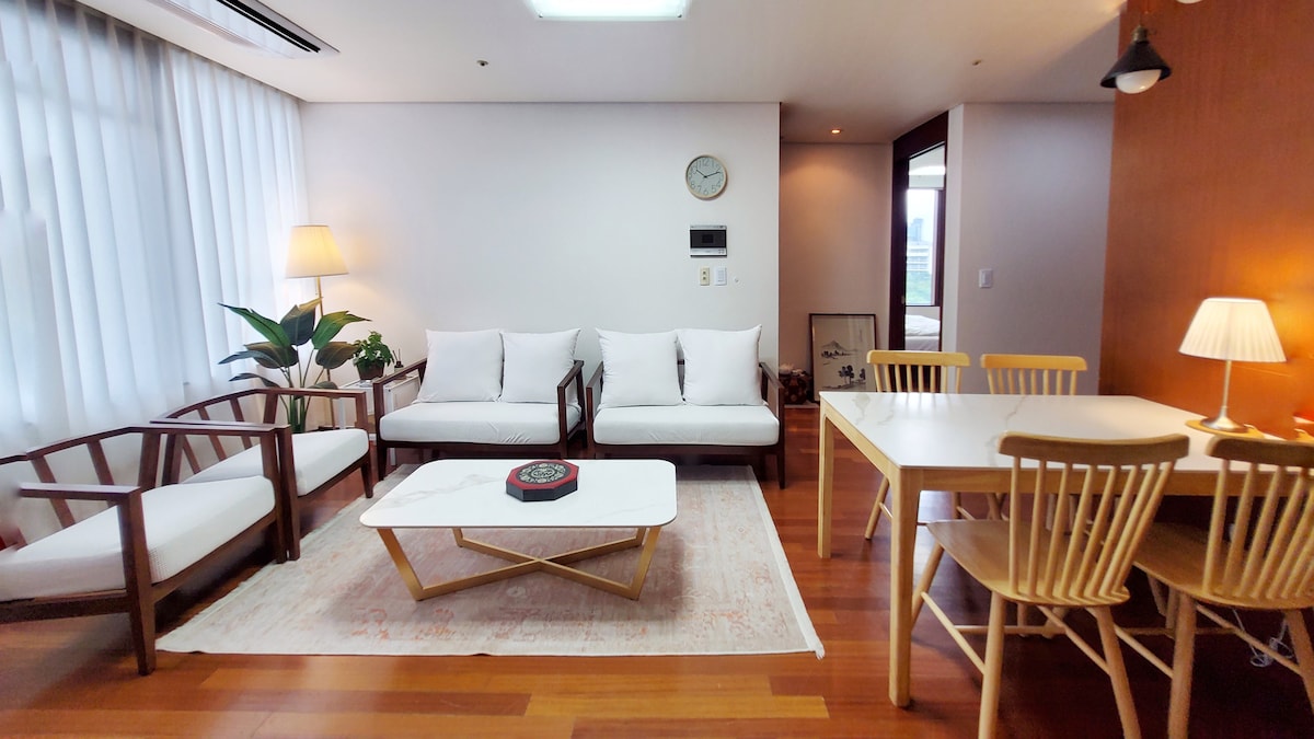 The living area features a spacious layout with two white sofas and a coffee table centrally positioned. A dining table with wooden chairs is visible beside a large window with sheer curtains. Indoor plants add a touch of greenery, complemented by warm lighting from floor and table lamps.