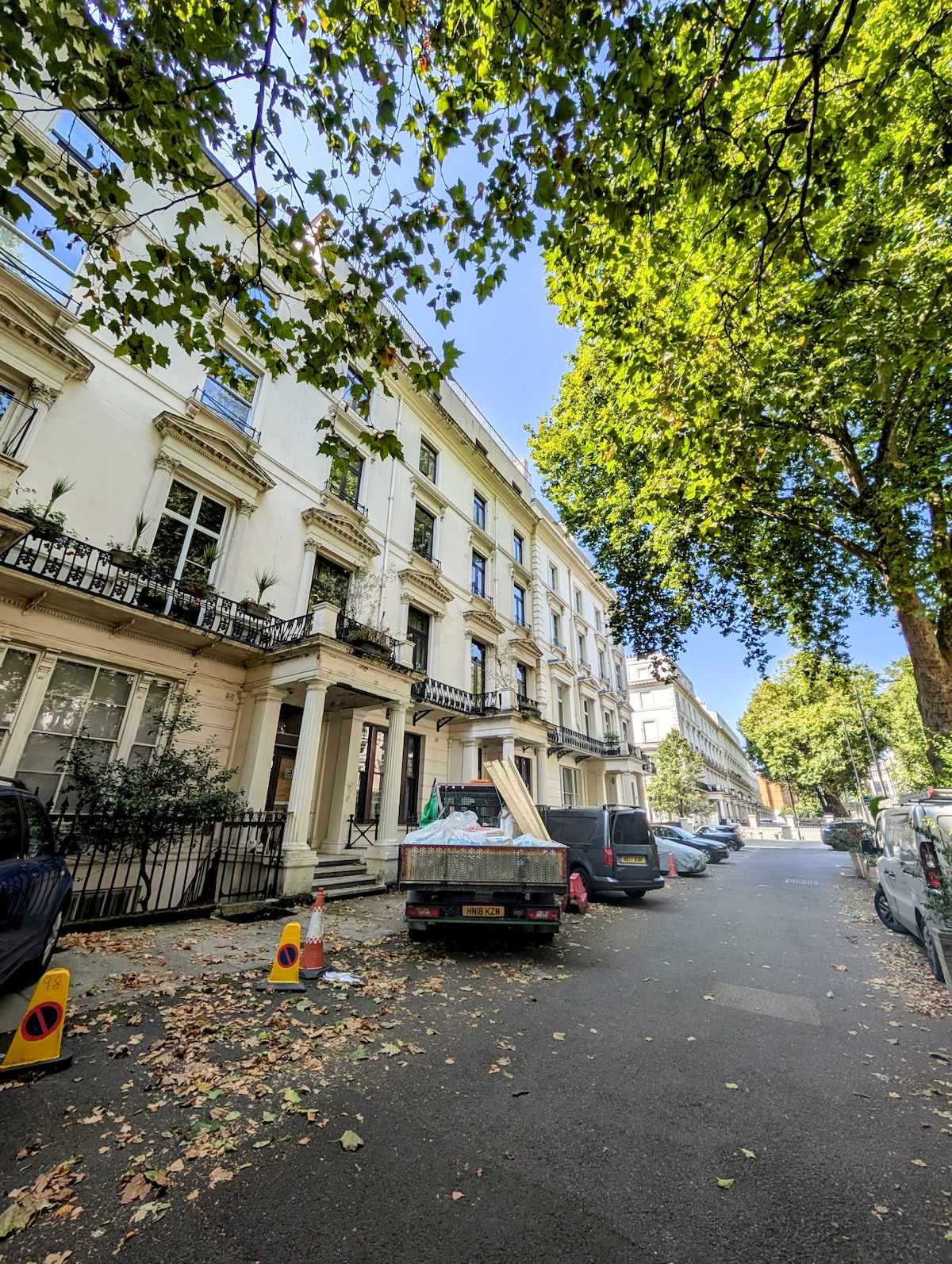 A leafy street lined with trees showcases a classic white building featuring balconies. Various parked cars are visible along the curb, with a truck unloading materials in front. Sunlight filters through the foliage, creating a serene ambiance.