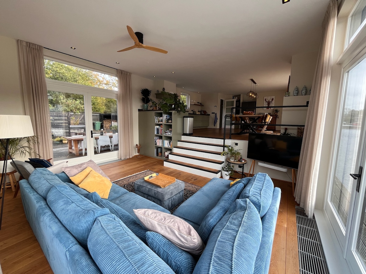 A spacious living area features a large blue sectional sofa and a coffee table, surrounded by natural light from expansive glass doors. A cozy rug lies beneath, and steps lead to an elevated dining area visible in the background, creating a welcoming atmosphere.