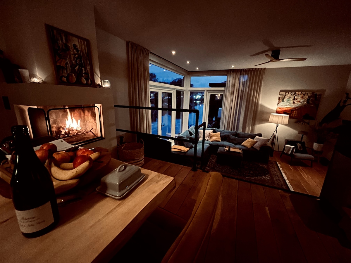A cozy living area is illuminated by warm light from a fireplace, with large windows revealing an evening view outside. A dining table is set with a bottle of wine and a bowl of apples, while comfortable seating arrangements invite relaxation.