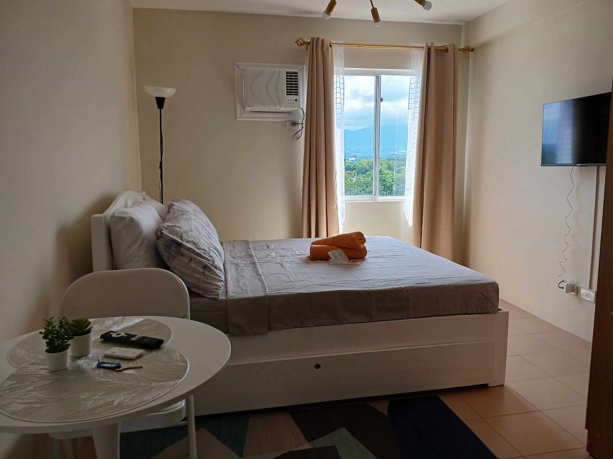 A cozy studio space features a comfortable double bed with neutral linens and an orange towel elegantly displayed. A small round table with two chairs is positioned nearby. Natural light enters through a large window, offering views of the mountains in the distance.