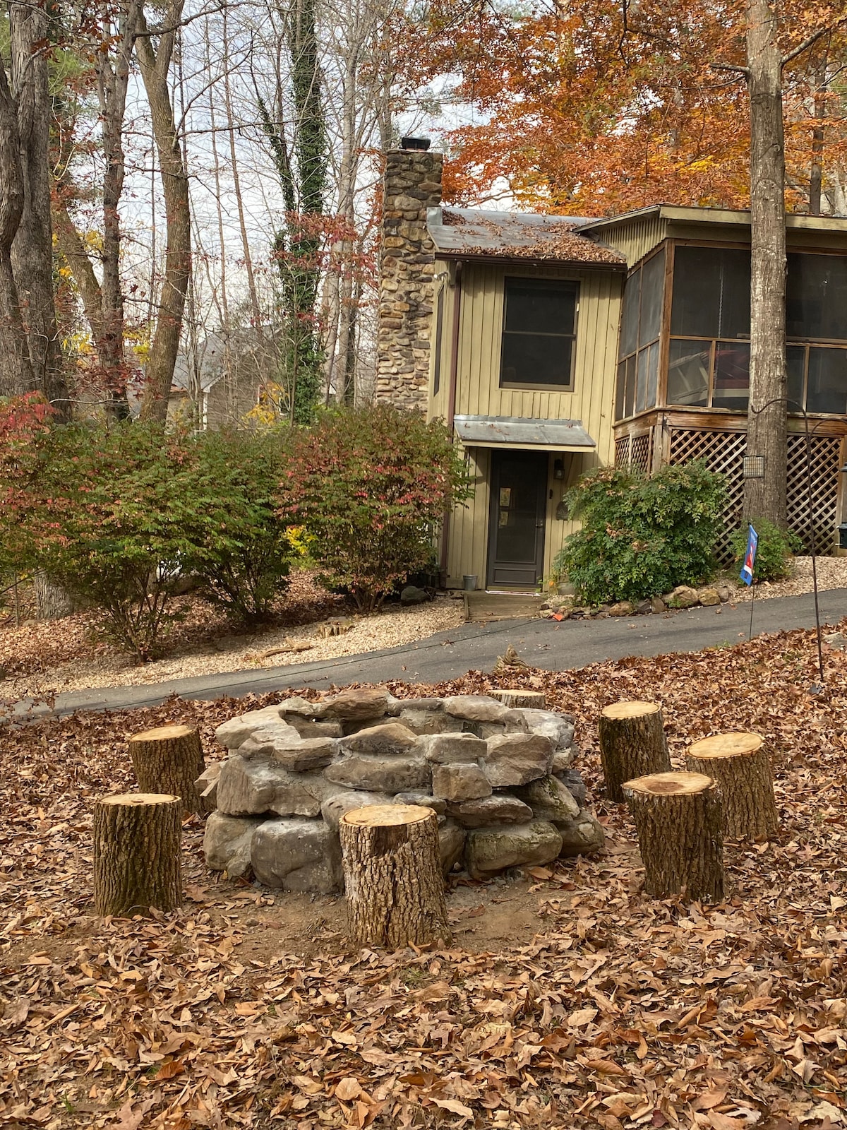 A rustic fire pit made of stone is surrounded by wooden stumps, creating a gathering space in a wooded area. The cabin is visible in the background, featuring a screened porch and a natural stone chimney, with autumn foliage enhancing the tranquil atmosphere.