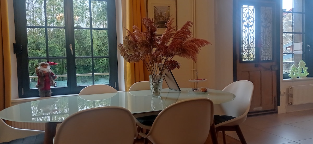 A dining area features a sleek, round table surrounded by white chairs. A glass vase with dried flowers sits at the center. Large windows with black frames allow natural light to fill the space, complementing the warm yellow curtains. A wooden door with decorative glass adds character.