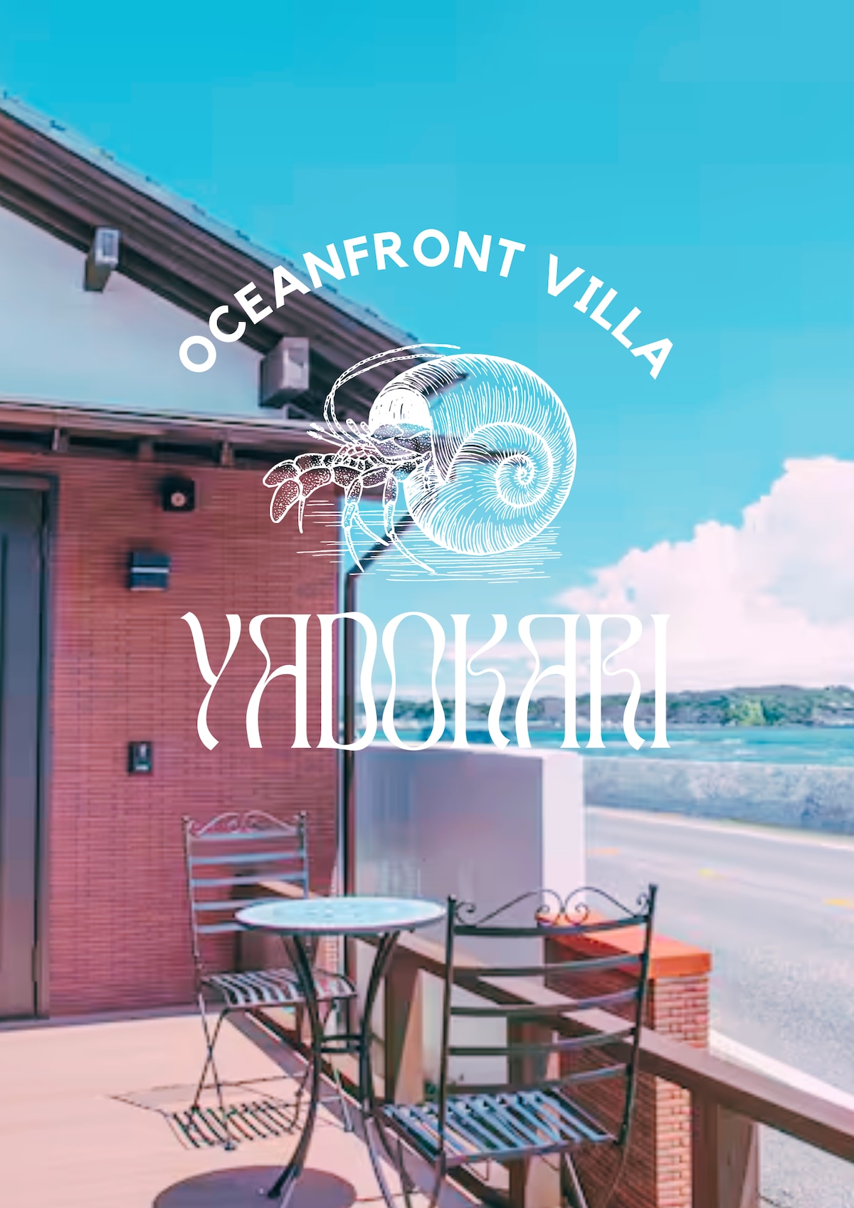 An outdoor patio area is presented with two metal chairs and a small round table, offering a view of the street and nearby surroundings. The sky is bright and clear, enhancing the inviting atmosphere of the sea-facing space.