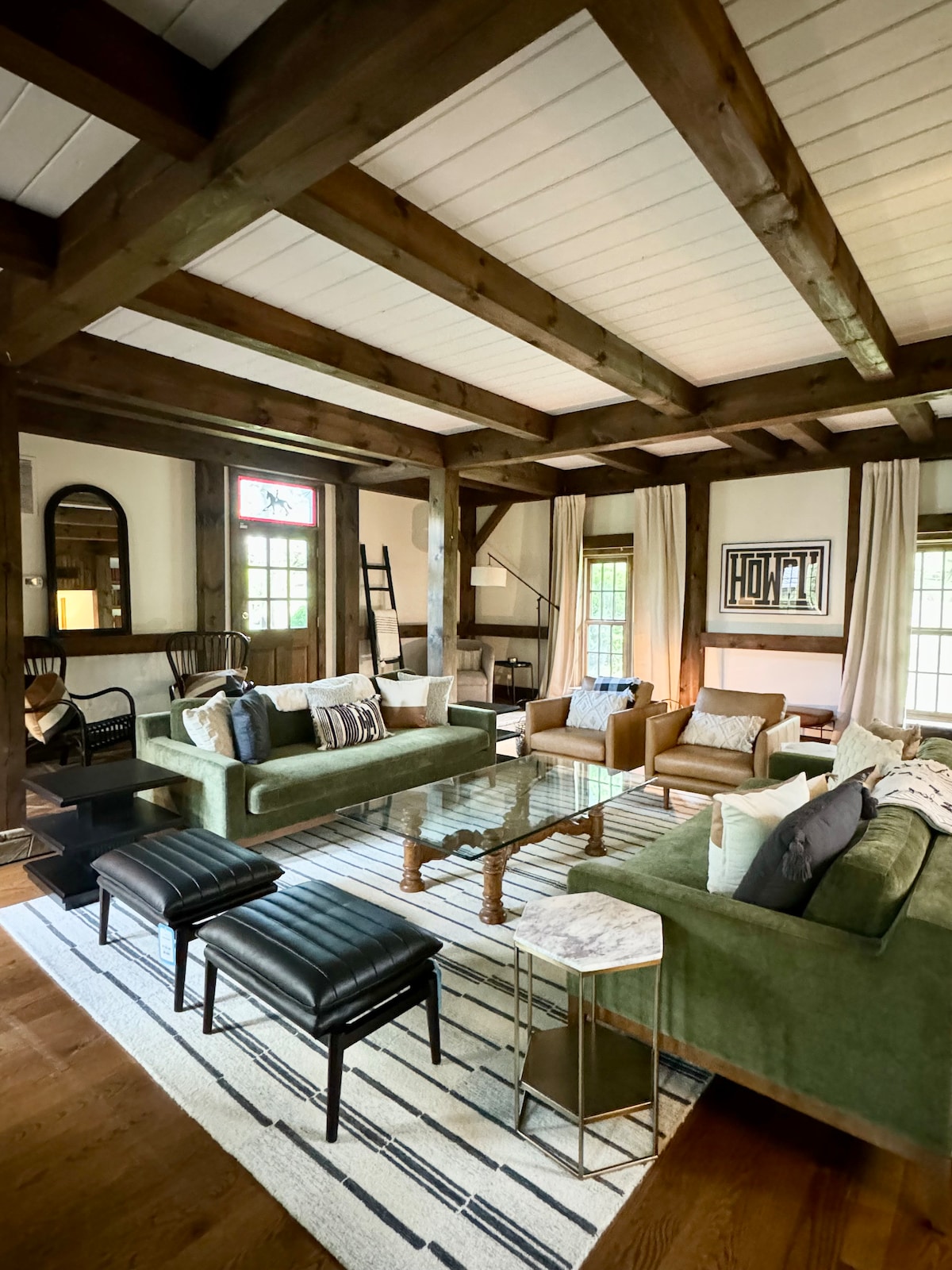 A spacious living area features exposed wooden beams and large windows. Comfortable seating is provided by green sofas and black benches, arranged around a glass coffee table. Soft textures and a striped area rug contribute to the inviting atmosphere.