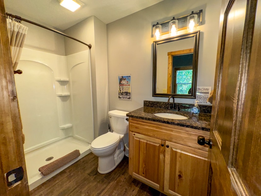 A well-appointed bathroom features a shower with a curved rod and a granite countertop sink. Natural wood elements complement the space, which is illuminated by overhead lighting. A large mirror is positioned above the sink, and a small decorative mat is placed in front of the shower.