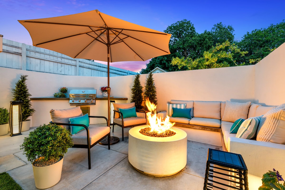 A private patio area features cozy seating around a circular fire pit, surrounded by decorative plants. An umbrella provides shade, while a built-in grill is positioned nearby. The soft glow of the fire offers a warm ambiance for outdoor relaxation.