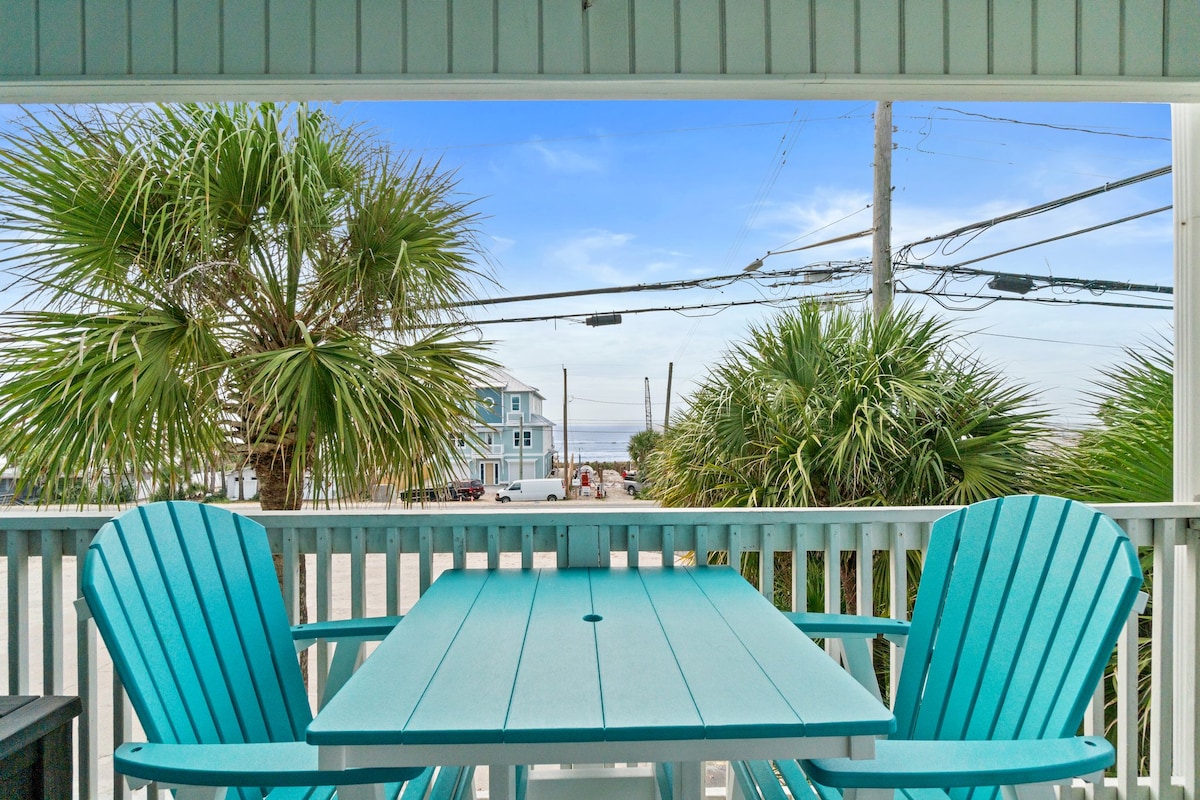 The balcony features a table and two teal adirondack chairs, inviting relaxation. Lush palm trees frame the view, providing a coastal atmosphere. A clear blue sky enhances the serene environment, while distant buildings hint at the nearby beach access.