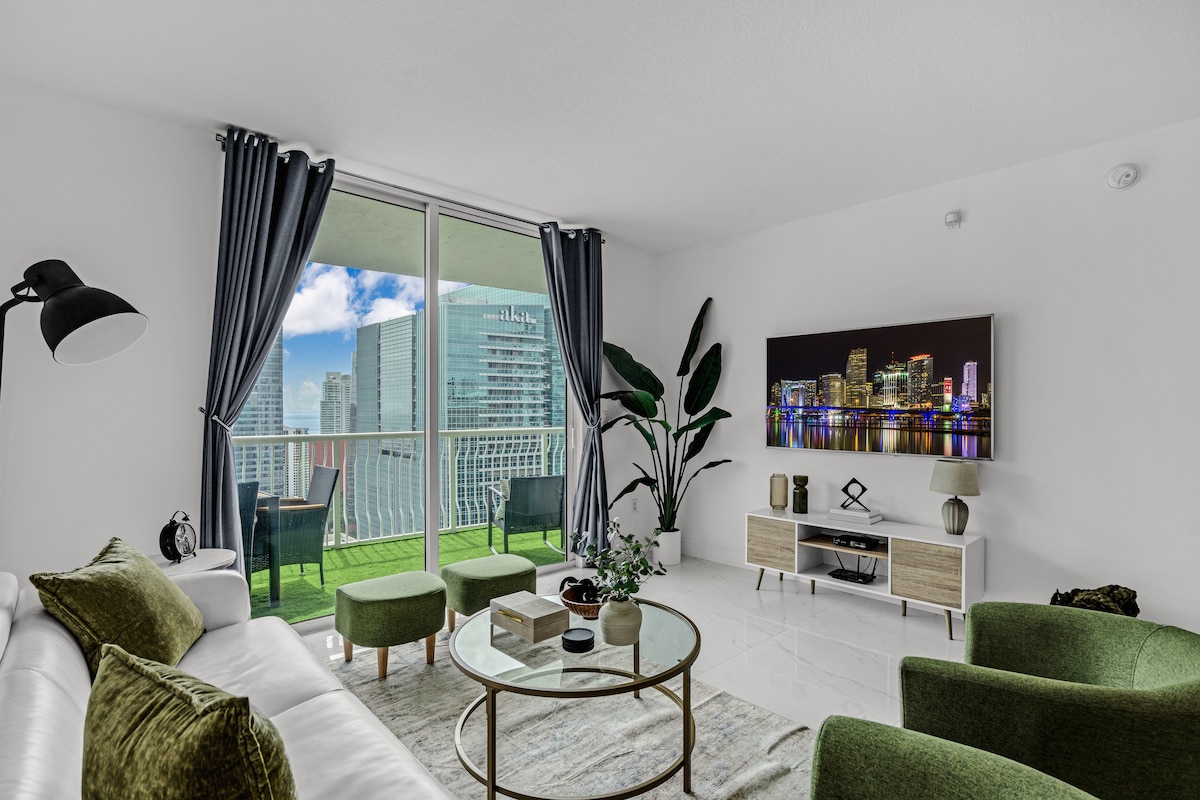 A modern living area features a plush white sofa complemented by green accent chairs. A round coffee table sits at its center. Floor-to-ceiling windows offer views of the cityscape and balcony access, while decorative plants add a touch of nature.