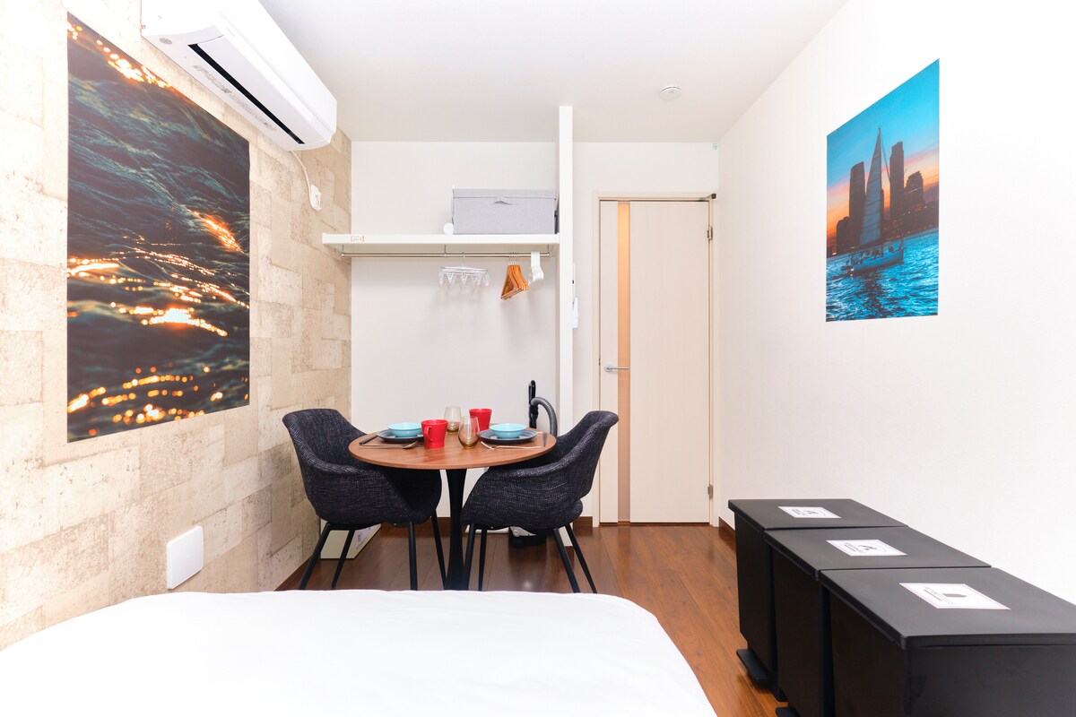 A cozy studio space features a comfortable bed with crisp white linens. A small dining area with two black chairs and a round table is seen, set with red cups. A scenic wall art piece adds character, while warm wood flooring completes the ambiance.
