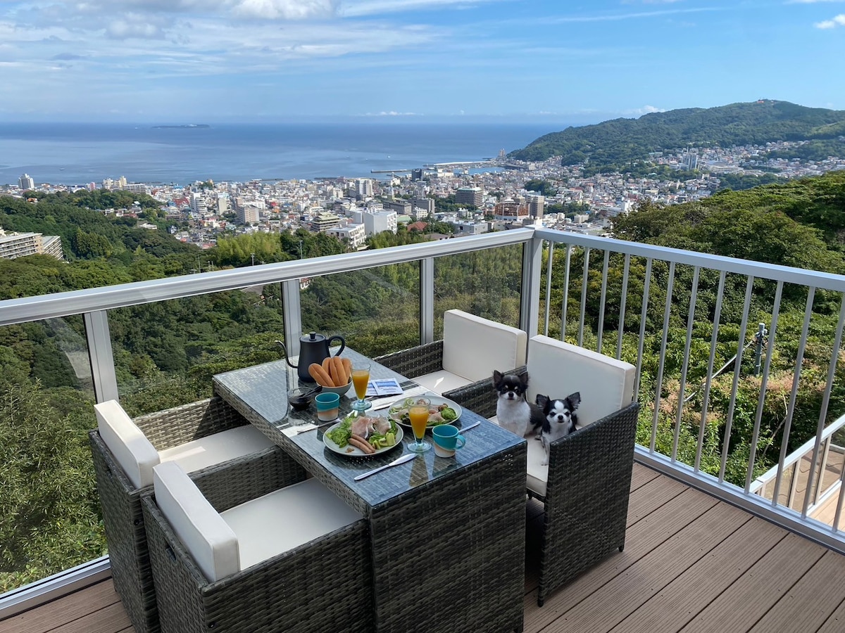 A glass-topped dining table is set for breakfast, surrounded by four wicker chairs. Two small dogs are seated at the table. The expansive ocean view and cityscape can be seen through the glass railing, enhancing the outdoor dining experience.