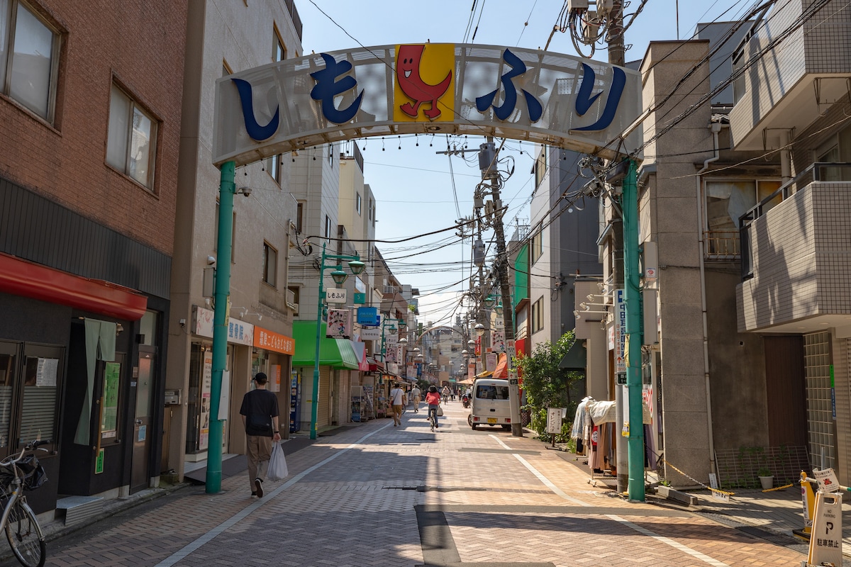 An inviting commercial street is presented, featuring an archway with colorful signage. The area is lined with shops and restaurants, while pedestrians stroll leisurely. Power lines and streetlights create a typical urban atmosphere, enhancing the accessibility and vibrancy of the location.