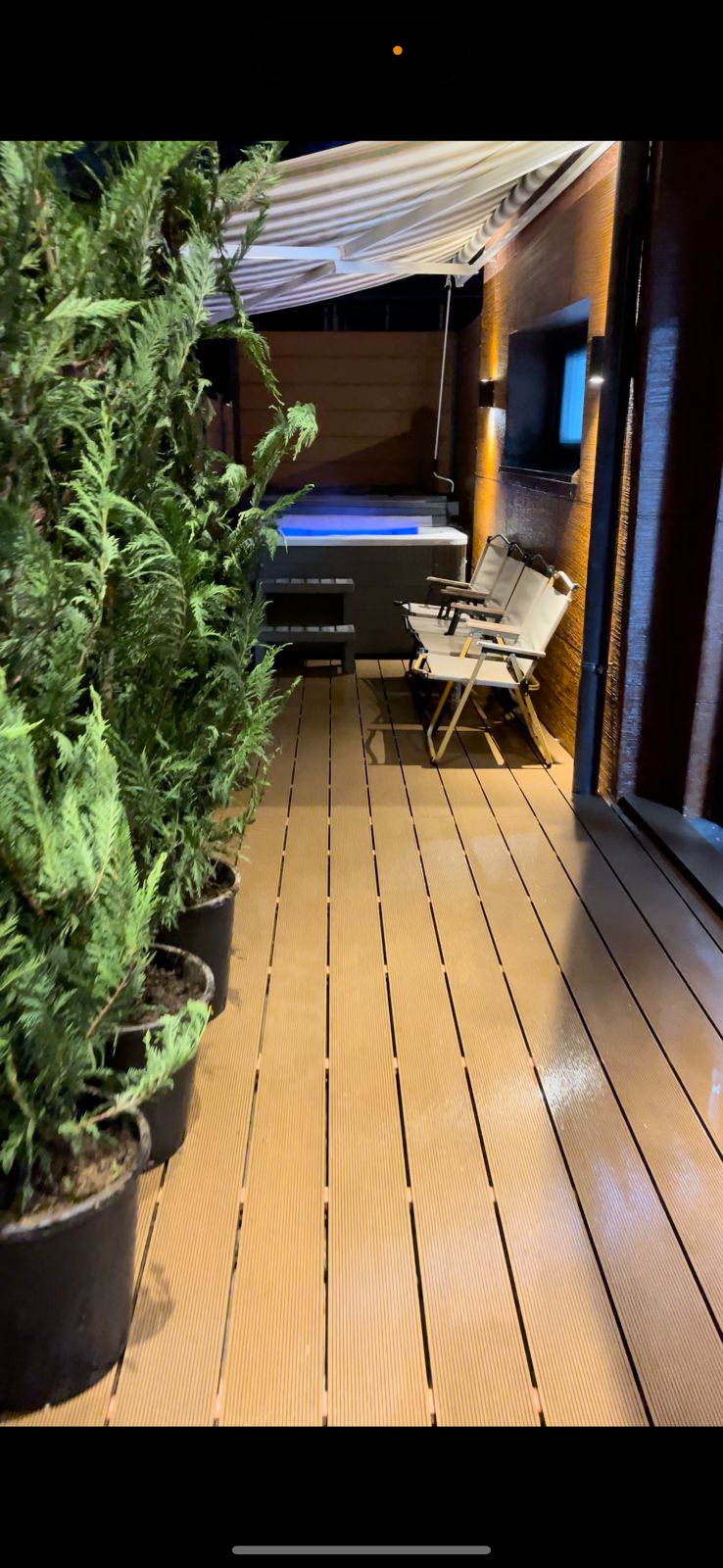 A well-maintained outdoor space features wooden decking bordered by potted green plants. Two chairs are positioned against a wall, alongside a hot tub designed for relaxation, under a shaded area created by a canopy.