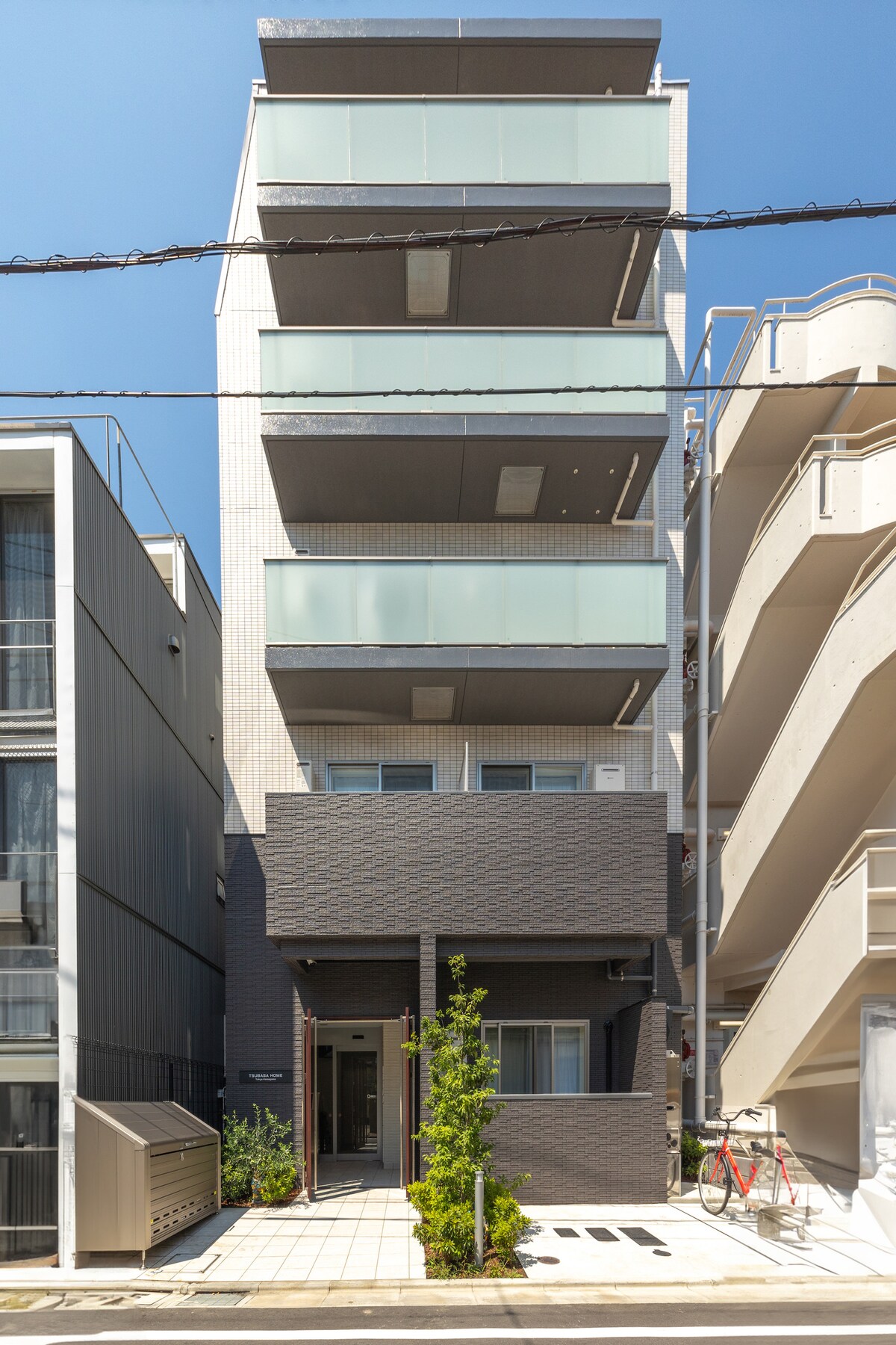 A modern four-story building features a sleek exterior with a mix of light and dark materials. Large glass balconies create a bright and open appearance, while the clean pathway leads to the entrance surrounded by minimal landscaping.