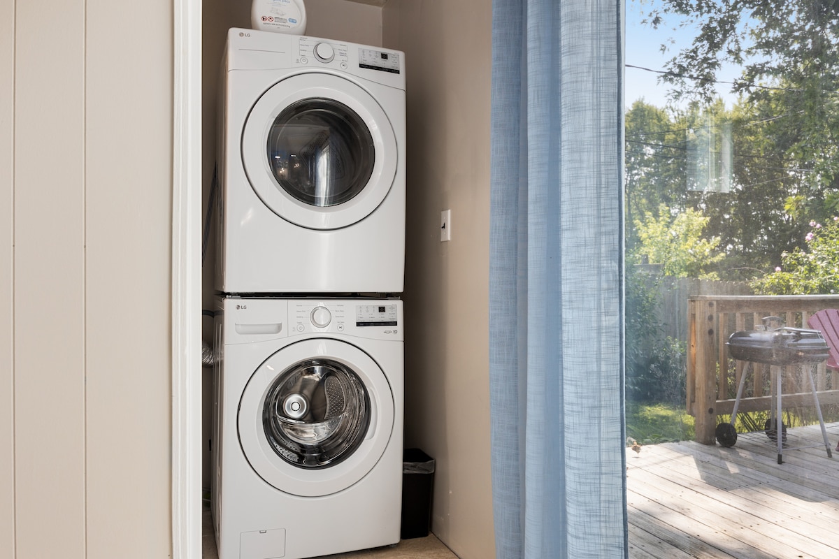 A stacked washer and dryer unit is positioned against a wall in a laundry area. Natural light filters through sheer curtains, suggesting a connection to the outdoor space beyond. A glimpse of a wooden deck and outdoor furniture can be seen through the nearby opening.
