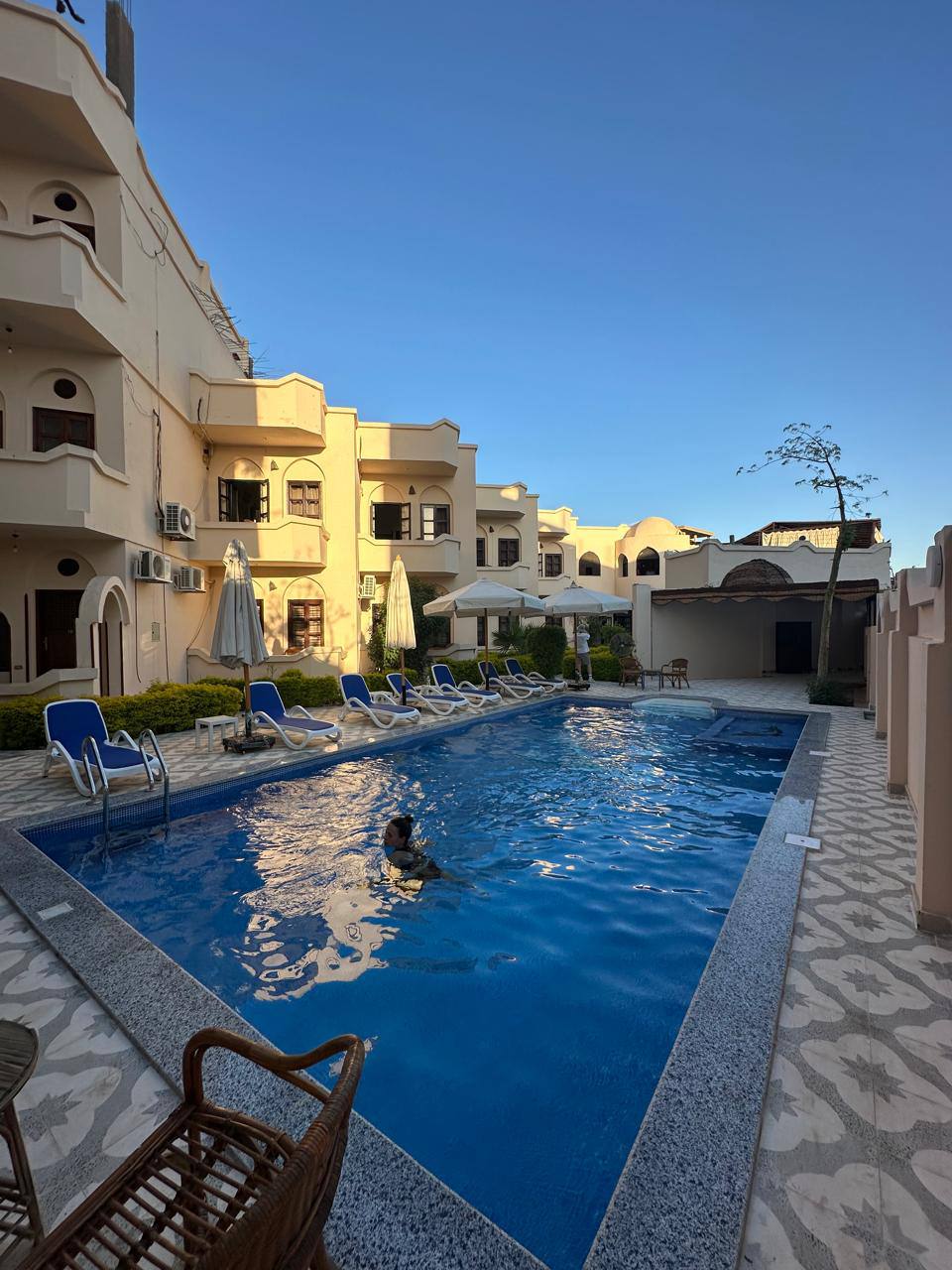 A serene swimming pool is surrounded by sun loungers and umbrellas, set within a courtyard of a multi-level building. Soft sunlight casts reflections on the water, while lush greenery lines the pool area. The scene conveys a relaxed and inviting atmosphere.