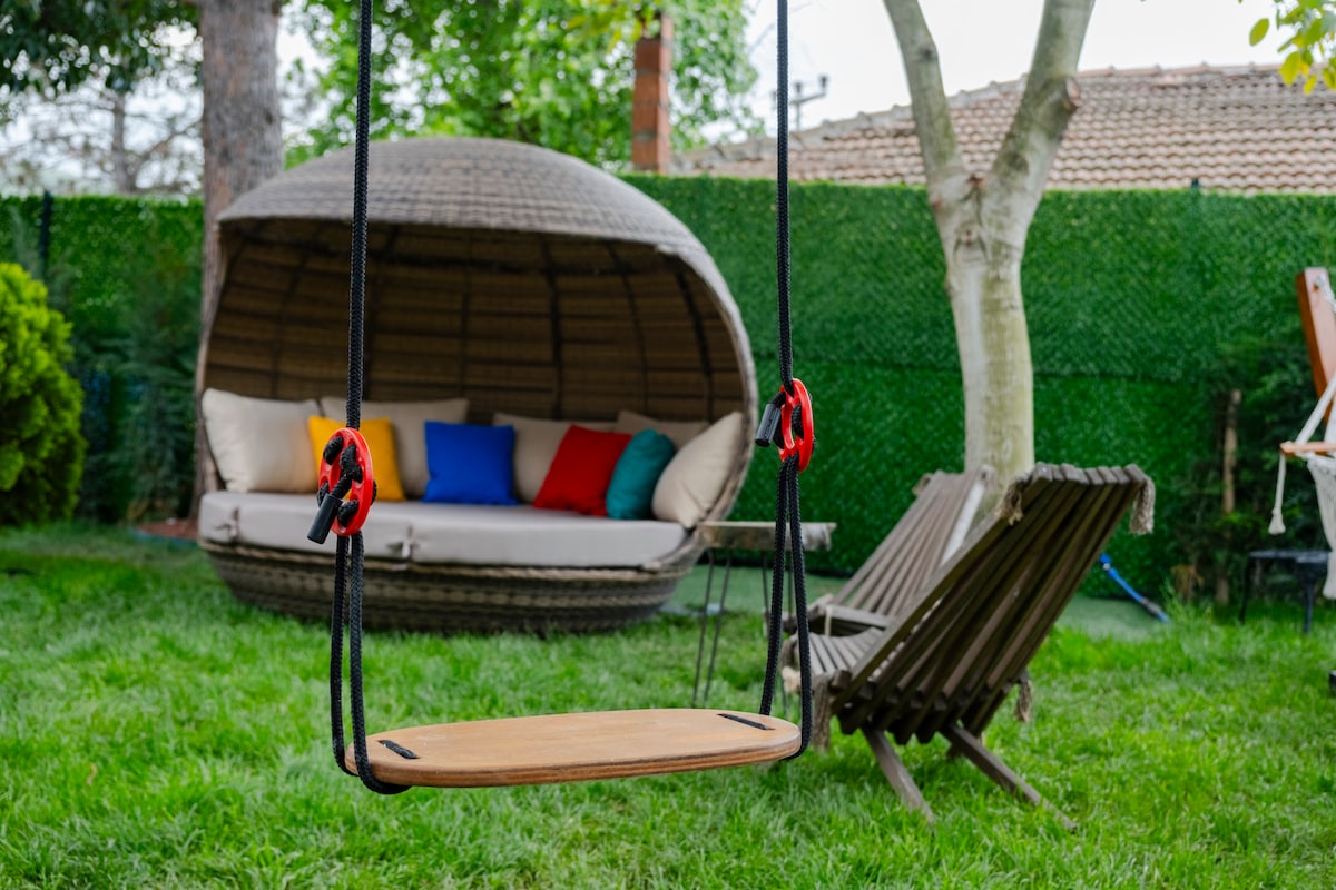 A backyard area features a wooden swing suspended from ropes, positioned on lush green grass. In the background, a cozy, semi-circular canopy bed with colorful cushions provides a comfortable lounging space. A wooden lounge chair is also visible, enhancing the outdoor relaxation area.