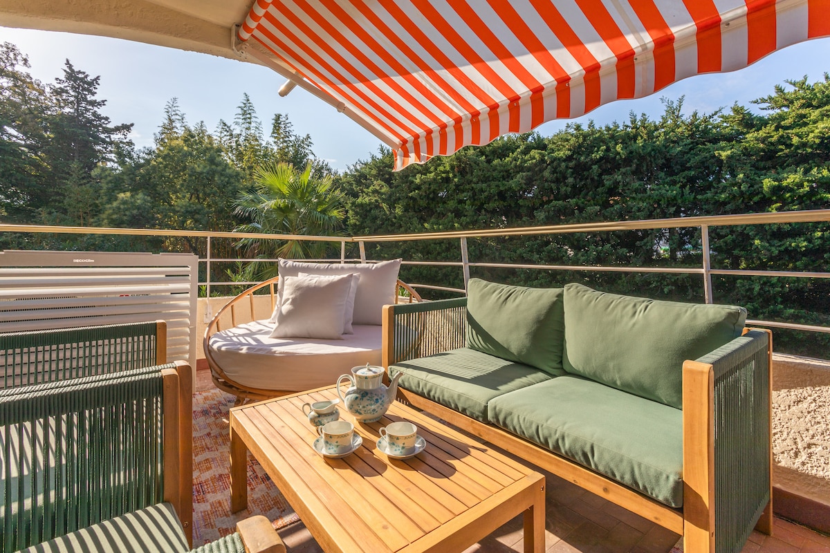 A spacious terrace features green seating arrangements and a wooden table set with tea cups. A shaded area is provided by a striped awning, while surrounding greenery offers privacy. A comfortable sunbed invites relaxation, creating an inviting outdoor space.