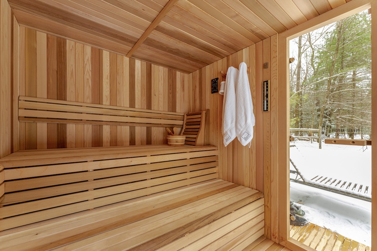 A wooden sauna interior features warm, smooth cedar walls and benches. Two white towels hang on a hook, while a wooden bucket sits on one of the benches. Sunlight illuminates the entrance, revealing a snowy landscape beyond.