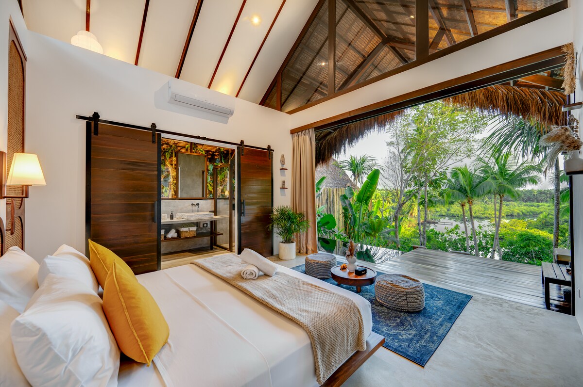 A spacious bedroom is designed with wooden sliding doors that open to a vibrant outdoor view. A plush bed with crisp linens is paired with decorative cushions, while a cozy seating area is complemented by a small table and lush greenery visible outside.