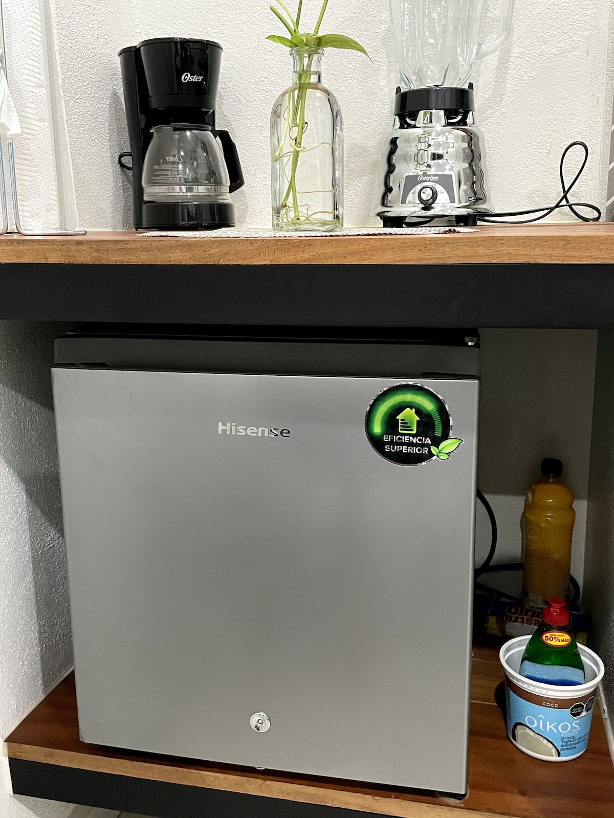 A small kitchenette shelf is displayed, featuring a Hisense mini-fridge, a coffee maker, and a blender. A bottle of yellow liquid and a decorative plant in a glass vase are also visible. A cup is seen along the bottom shelf.