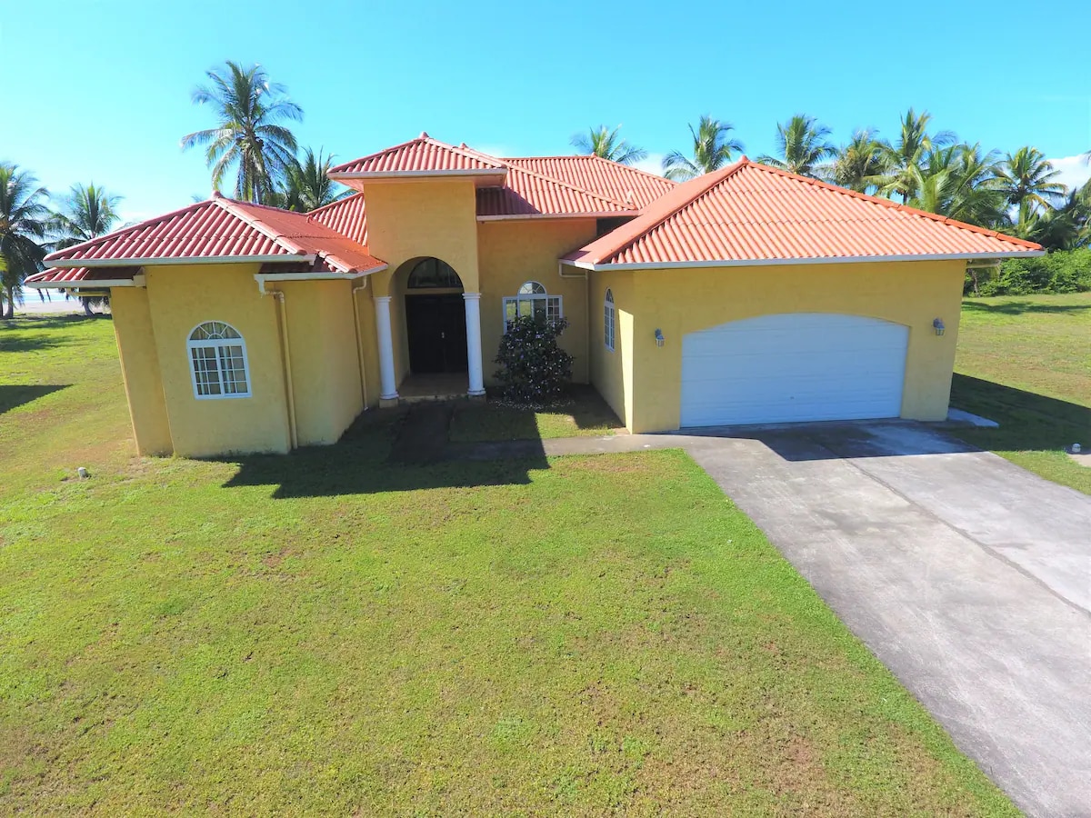 A vibrant villa with a red-tiled roof and yellow exterior is shown against a clear blue sky. Lush green grass surrounds the property, and palm trees are visible in the background. The garage is accessible from a paved driveway leading to the entrance.