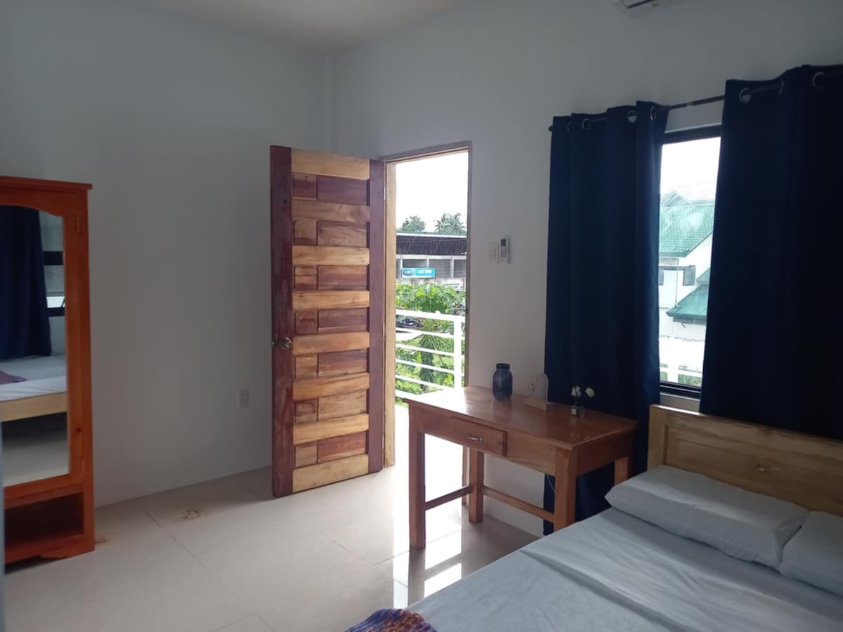 A well-lit room features a spacious bed next to a wooden table. Natural light enters through the large window, which is dressed with dark curtains. A wooden door leads outside, and a mirror is seen in the corner, enhancing the room's overall brightness.