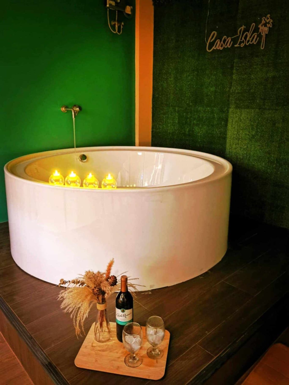A round bathtub is positioned on a wooden platform, illuminated by soft yellow lights. An arrangement of dried flowers and a bottle of wine sit on a wooden tray beside two glasses, against a backdrop of green walls and textured paneling.