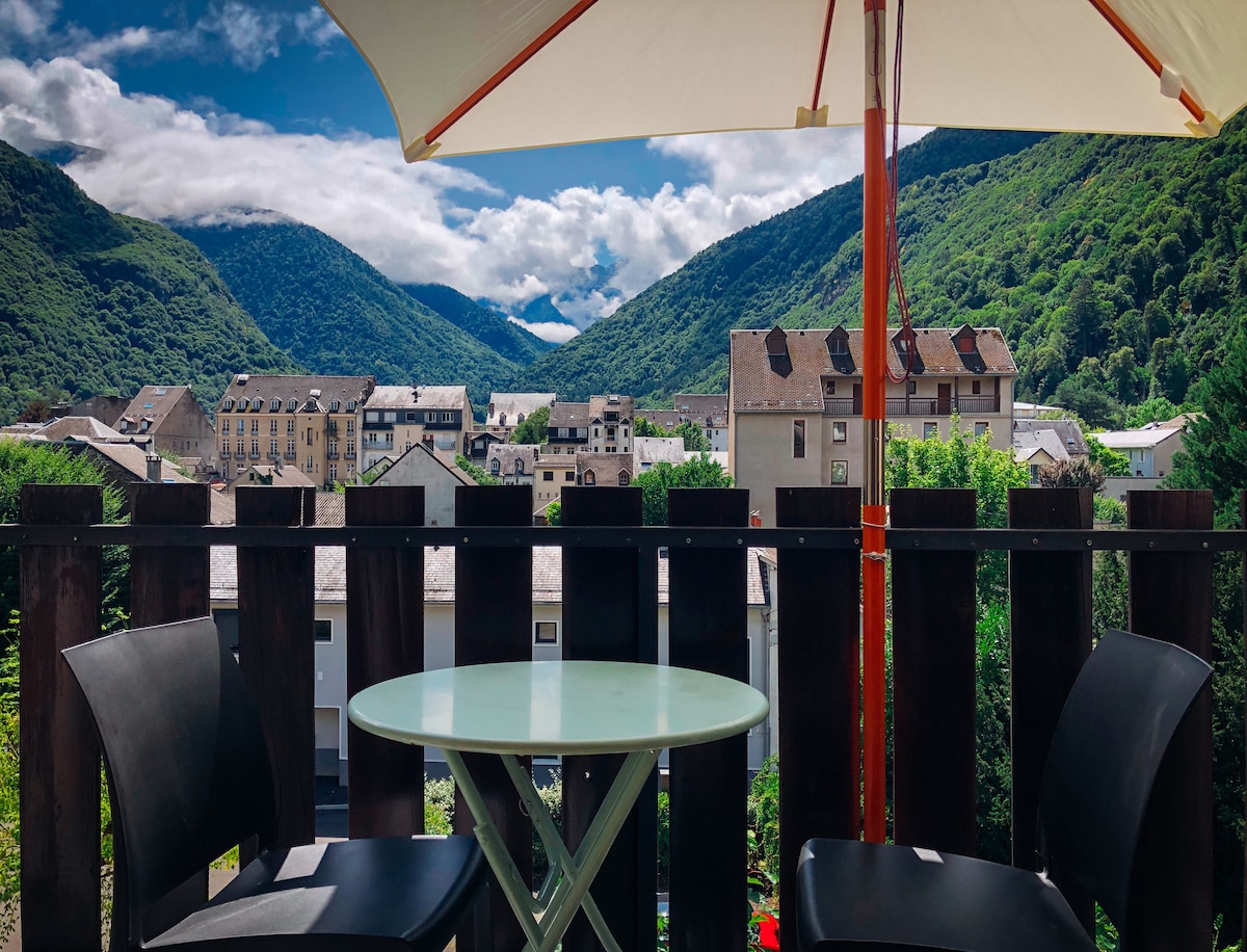 A terrace is depicted with a round table and two black chairs, overlooking lush green mountains and a scenic village. A large umbrella provides shade, framing the stunning view of the landscape under a partly cloudy sky.